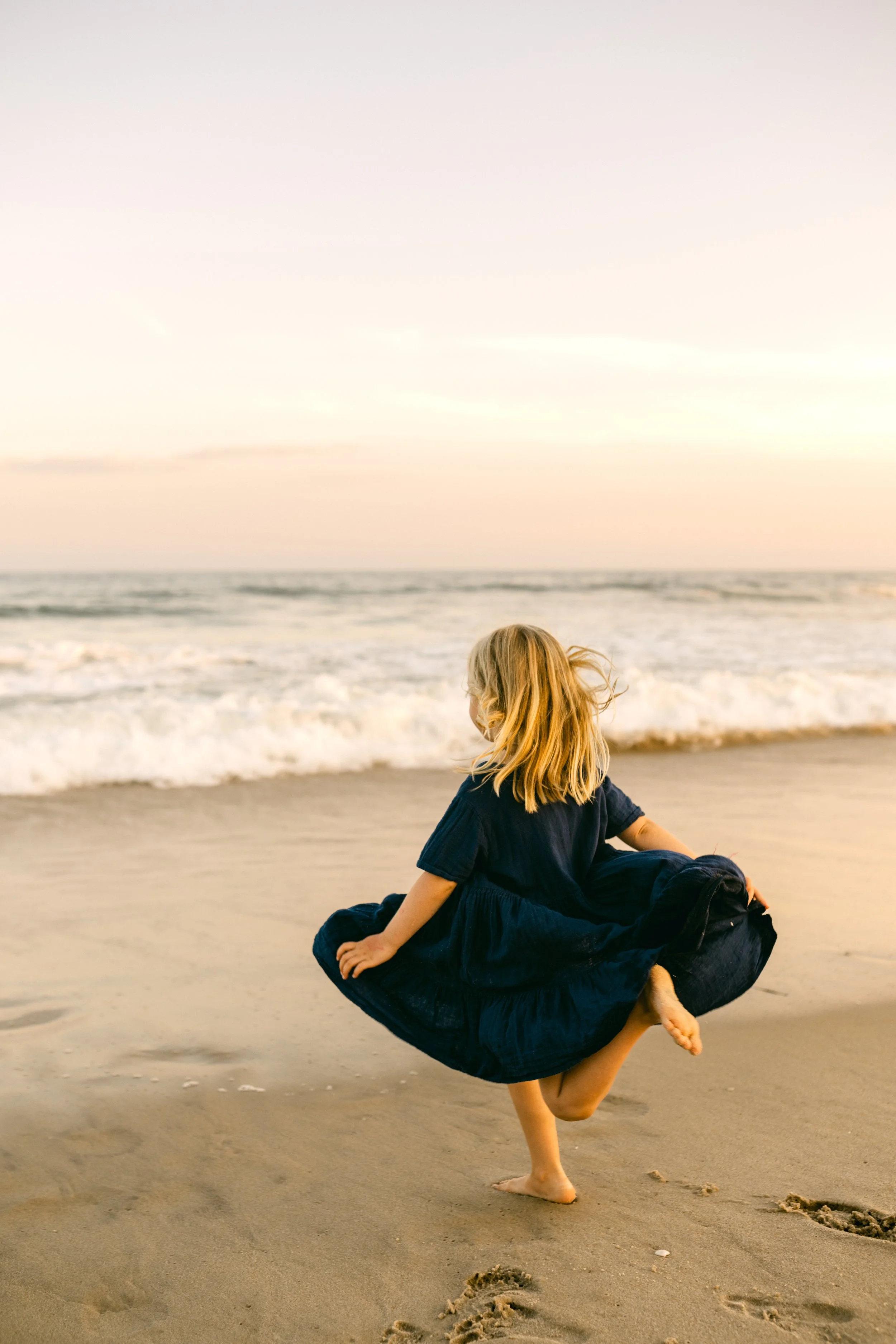 A young girl in a dark blue dress playing on a sandy beach near the ocean at sunset.