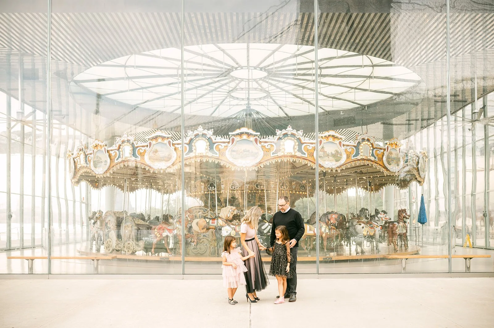 Family of four standing in front of a carousel inside a glass-enclosed pavilion, with the carousel horses visible through the glass.