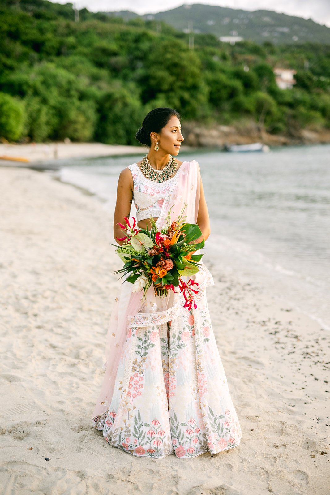 A woman in a floral dress and traditional jewelry holding a bouquet on a beach with greenery and hills in the background.