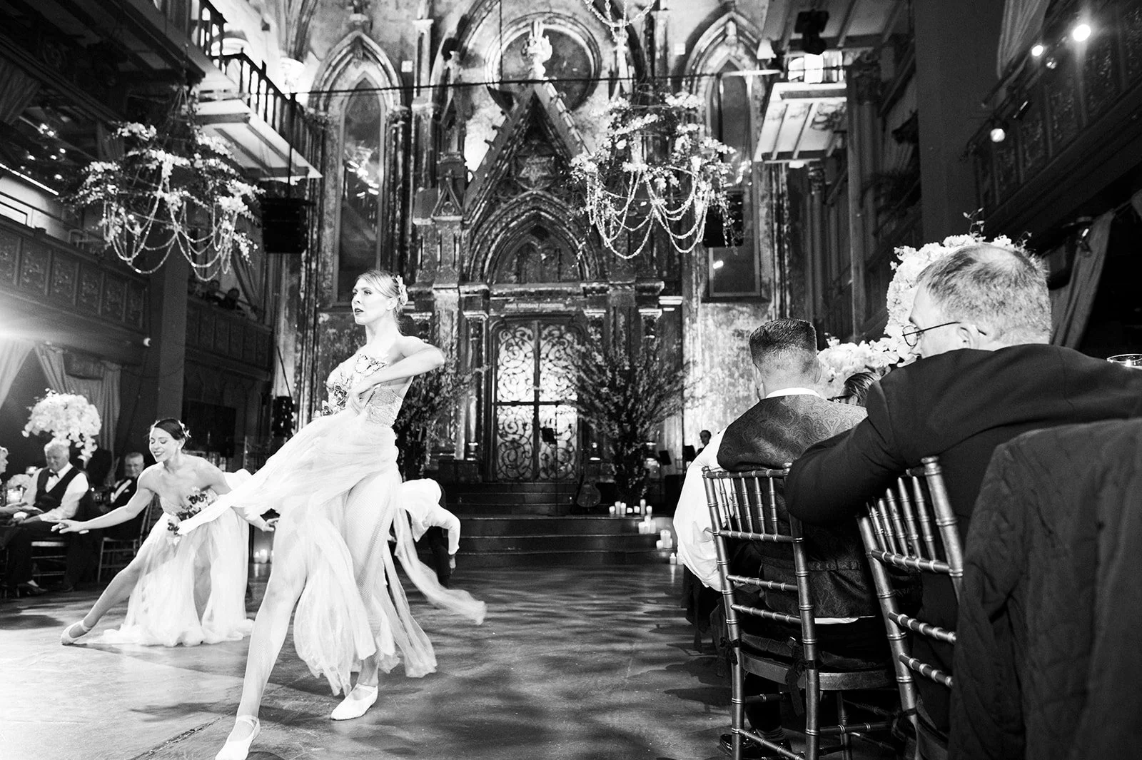Ballet dancers perform at a formal event in an elegant, ornately decorated hall with chandeliers and gothic architecture. Guests seated at tables watch the performance.