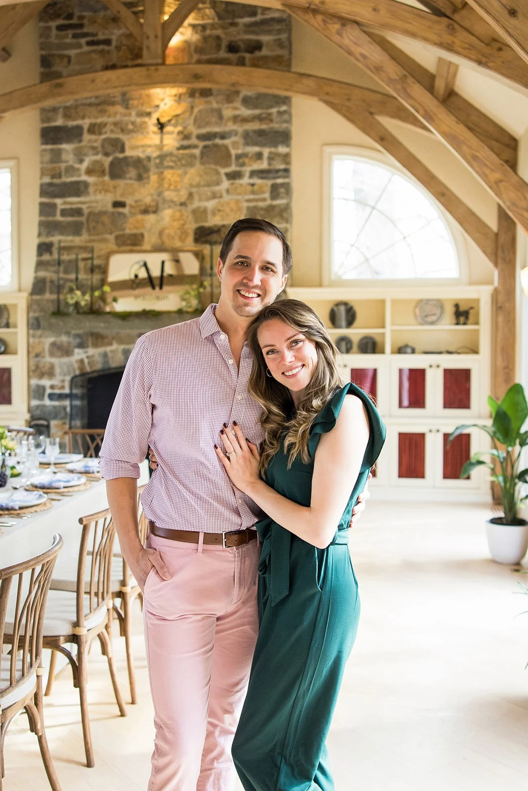 A smiling couple standing close together in a warmly lit, rustic-style dining or living room with wooden beams, a stone fireplace, and large windows, celebrating together.