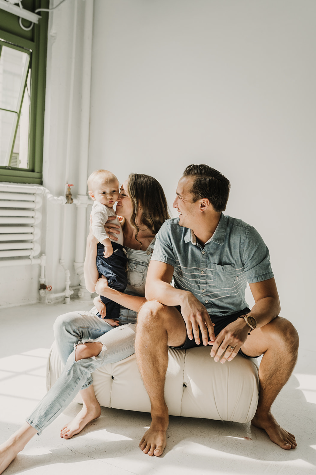 A family of three sitting together on a white bean bag in a bright room, with the mother holding a young child and the father smiling at them.