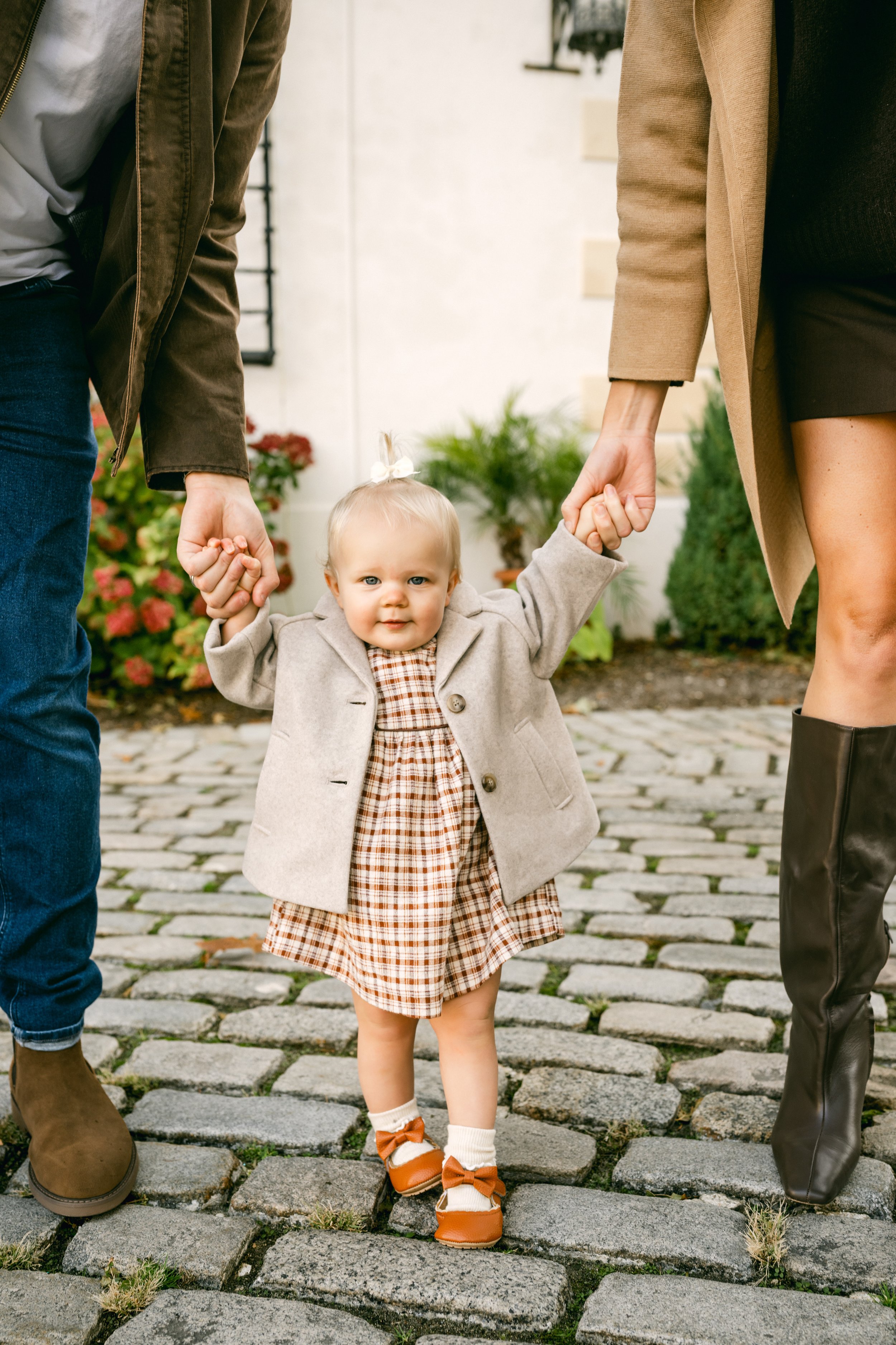 A young girl in a checked dress and beige coat holding hands with an adult on each side, standing on a cobblestone path outside with flowers and foliage in the background.
