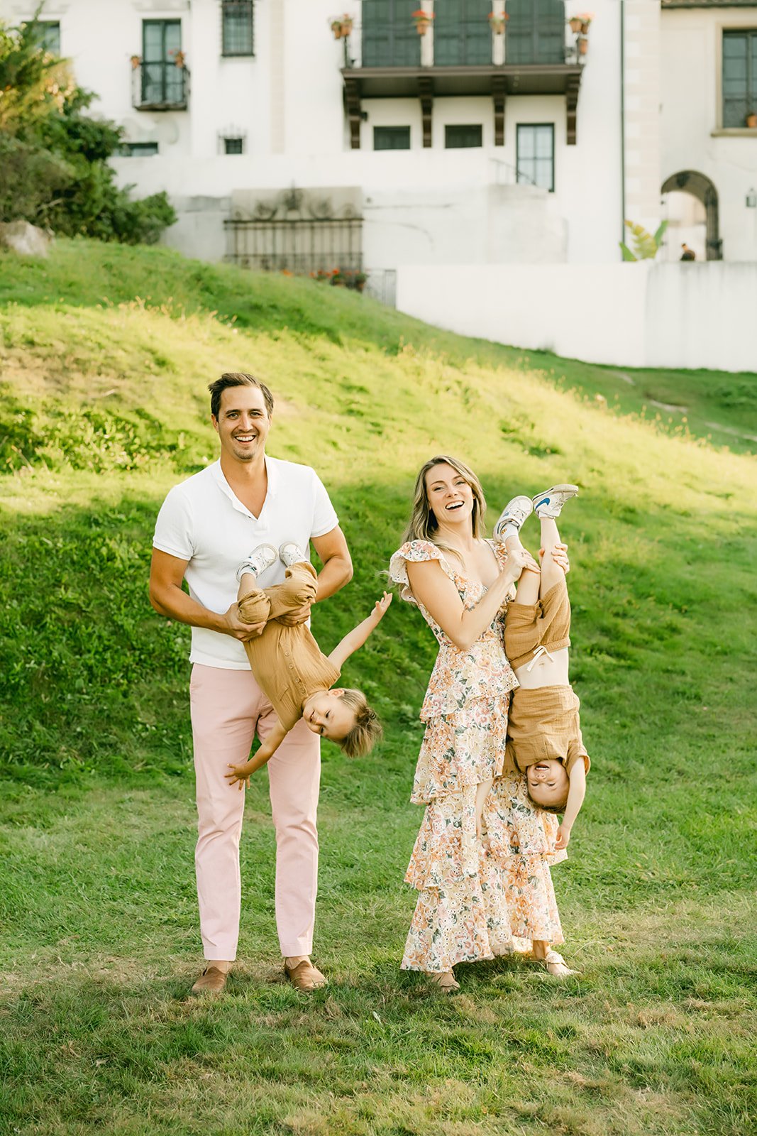 A family of four standing outdoors on a grassy hill, with two parents and two children. The parents are holding the children upside down, and all are smiling and laughing. The father is wearing a white polo shirt and pink pants, the mother is wearing a floral dress, and the children are dressed in brown tops and beige pants.