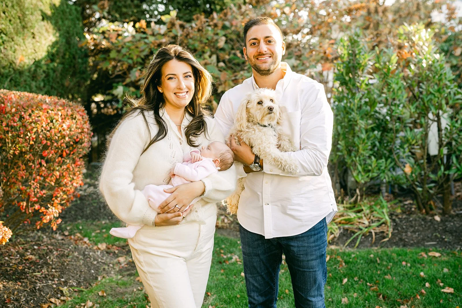 A smiling family of four standing outdoors in a park-like setting with bushes and trees with autumn foliage. The woman is holding a baby, and the man is holding a small dog. They are dressed in light-colored and casual clothes.