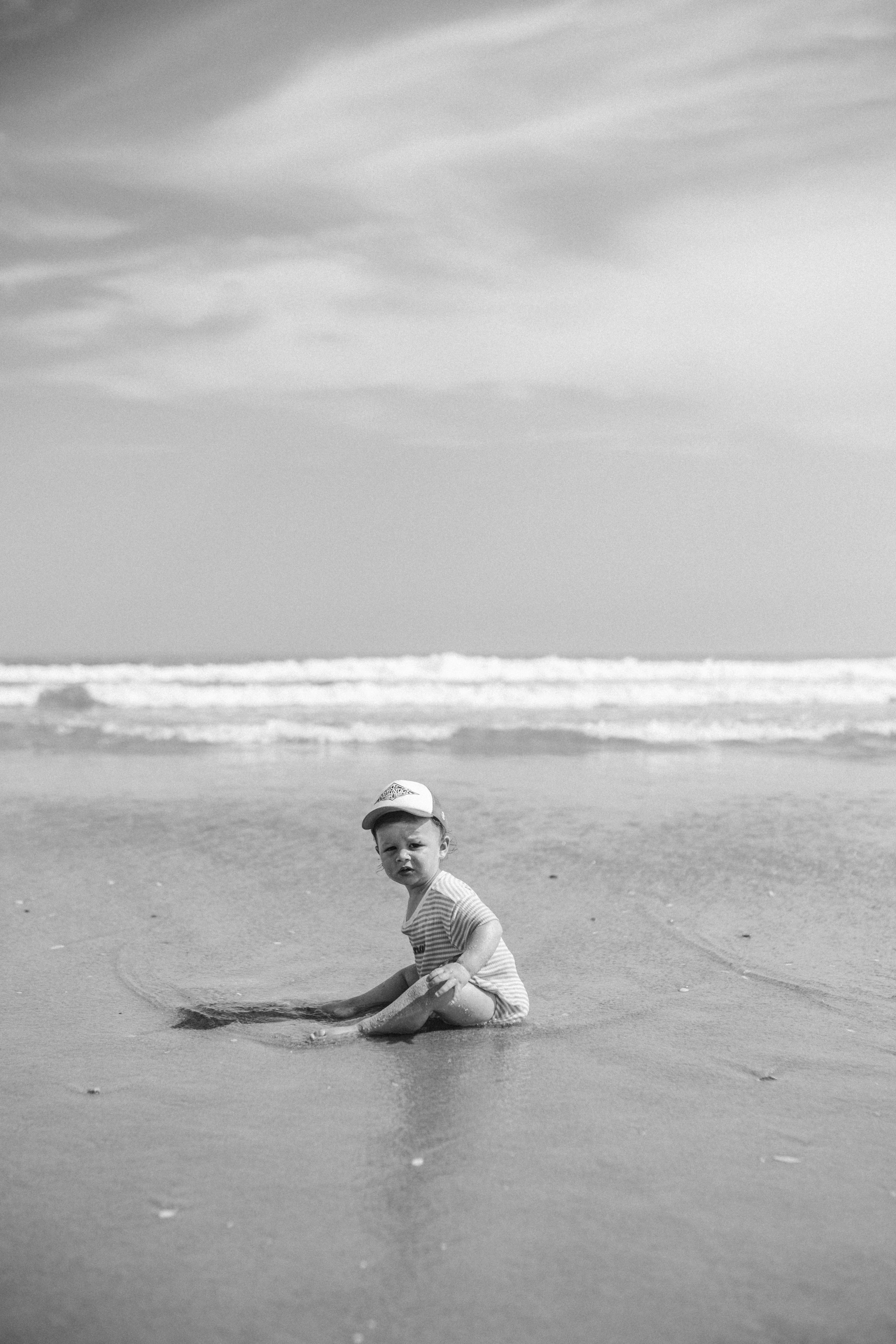A young child sitting in the sand on a beach, wearing a baseball cap and striped shirt, with the ocean and cloudy sky in the background.