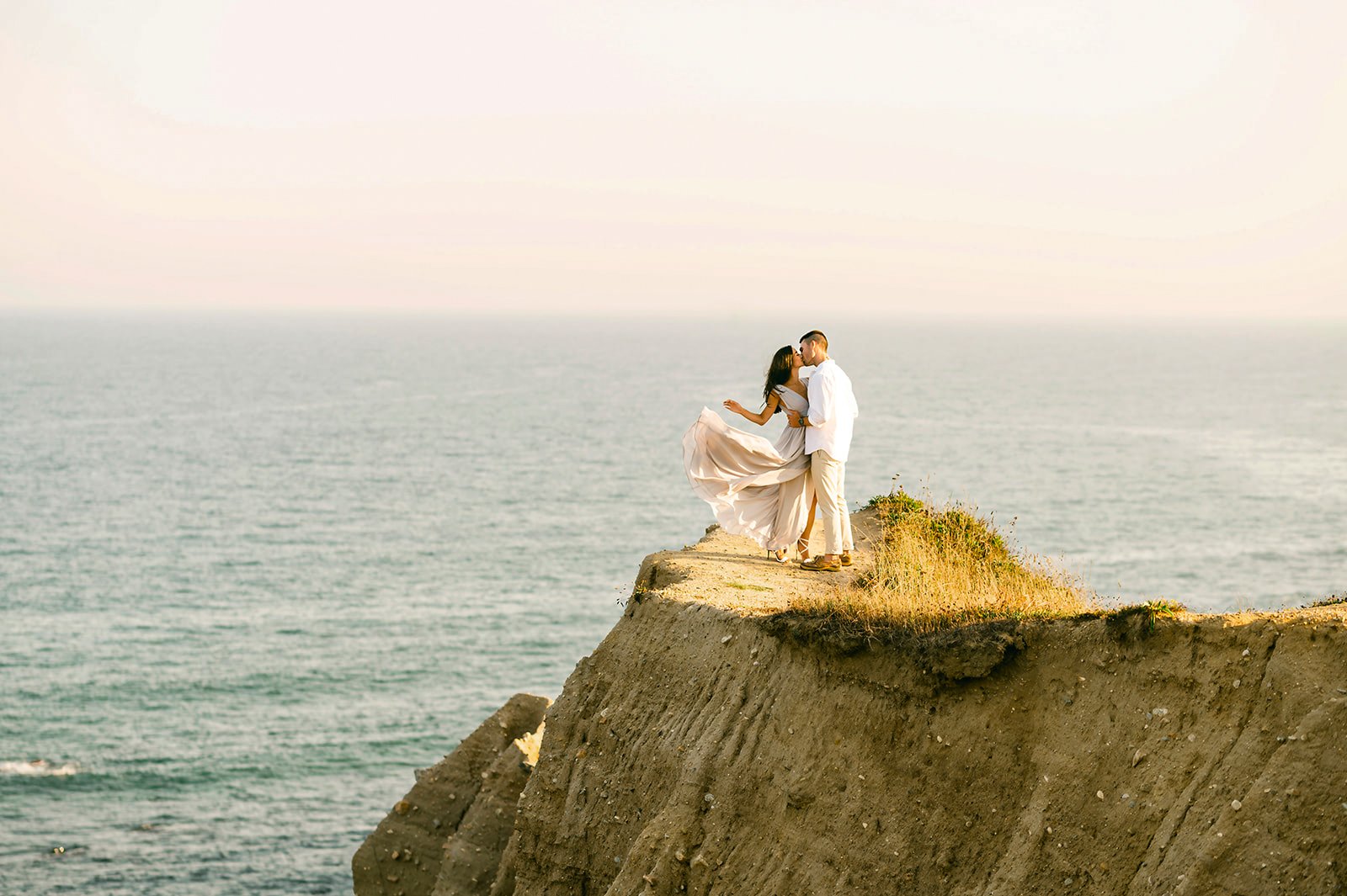 A couple is embracing and kissing on the edge of a cliff overlooking the ocean at sunset.