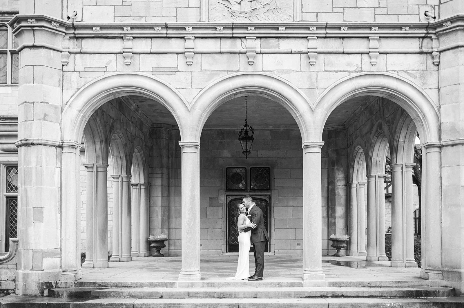 A black-and-white photograph of a newlywed couple standing under three arches in a historic stone building, with a chandelier hanging above them and ornate windows in the background.