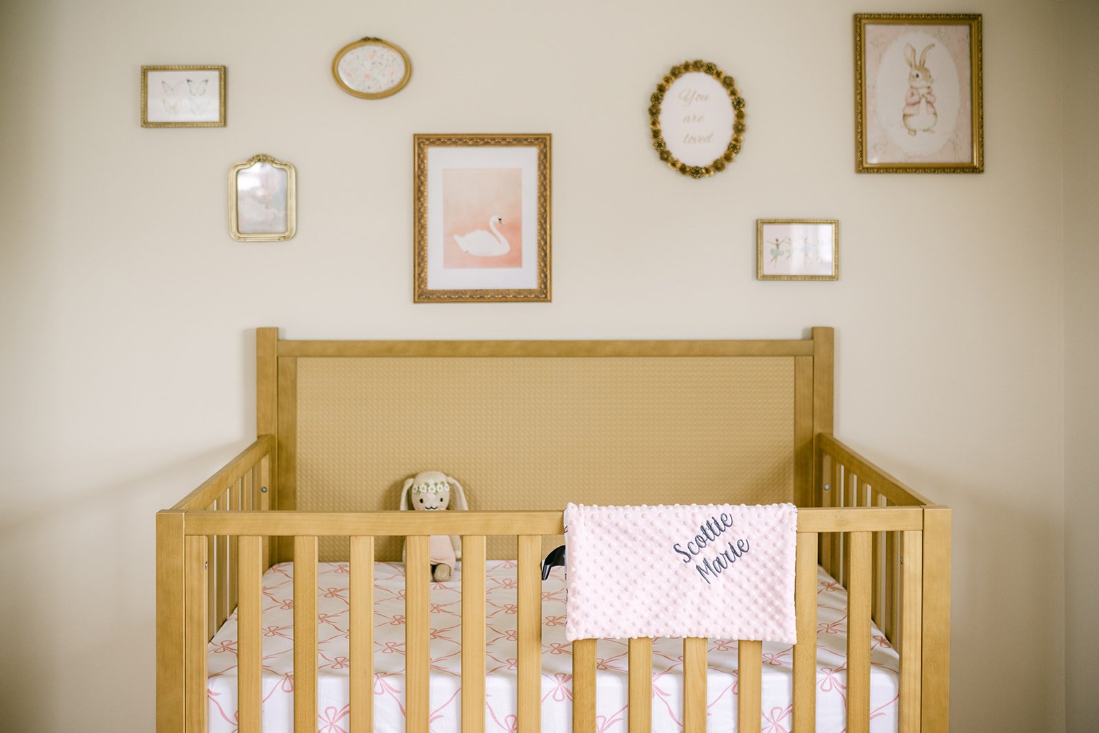 A wooden crib with a plush sheep toy and a pink blanket with the words "Scottie Mae" in cursive, against a wall decorated with framed pictures and artwork.
