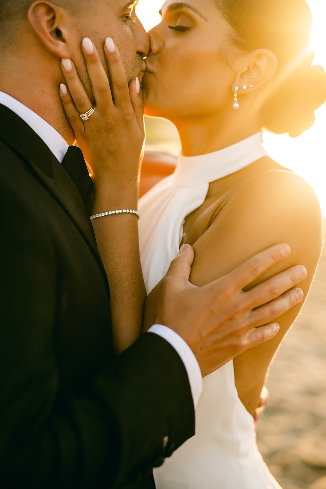 A couple sharing a romantic kiss during sunset, with the bride wearing earrings and a white dress, and the groom in a black tuxedo.