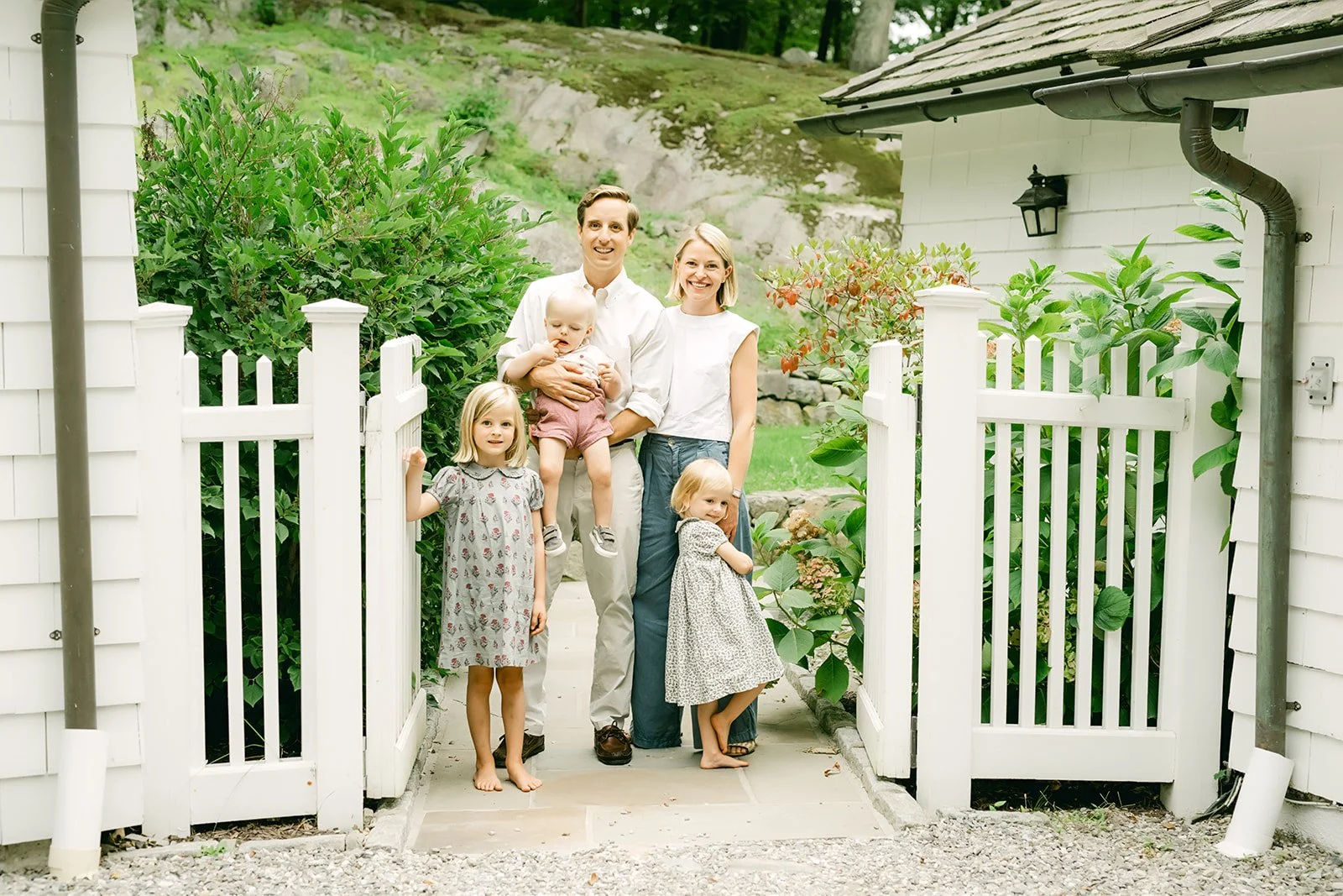A family of five standing at a white picket gate in front of a garden with green bushes and a rocky hillside behind their home.