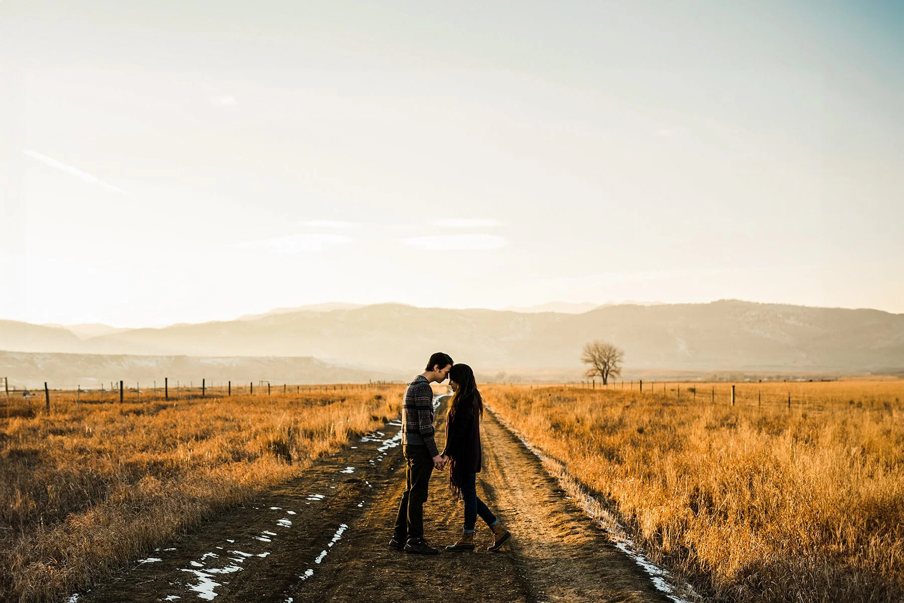 A couple holding hands and touching foreheads on a dirt road in a rural field during sunset or sunrise with distant mountains and a bare tree.