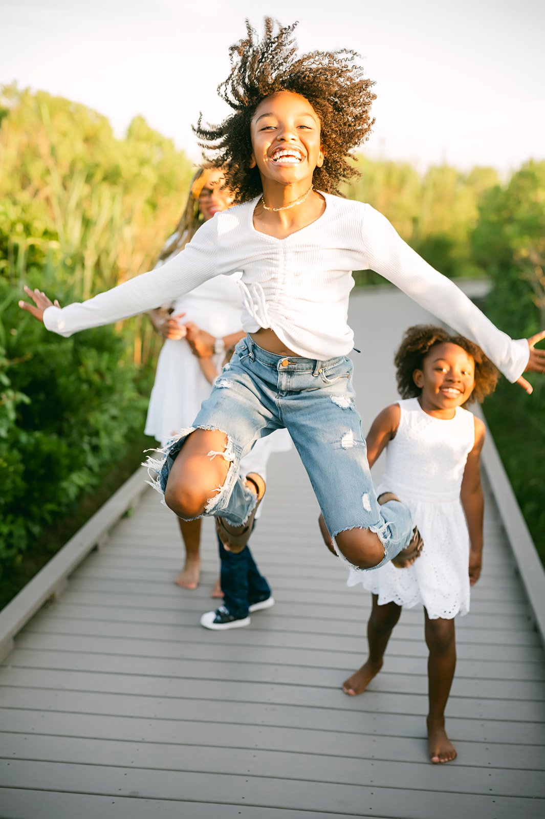 Four children, two girls and two boys, playing and jumping on a bridge outdoors, smiling, with green foliage in the background.