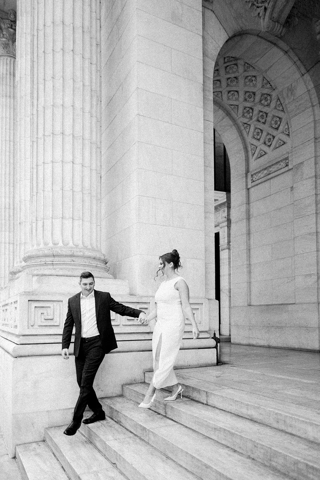 A man and woman in formal attire holding hands while stepping down stairs outside a classical building with large columns and detailed architecture, in black and white.