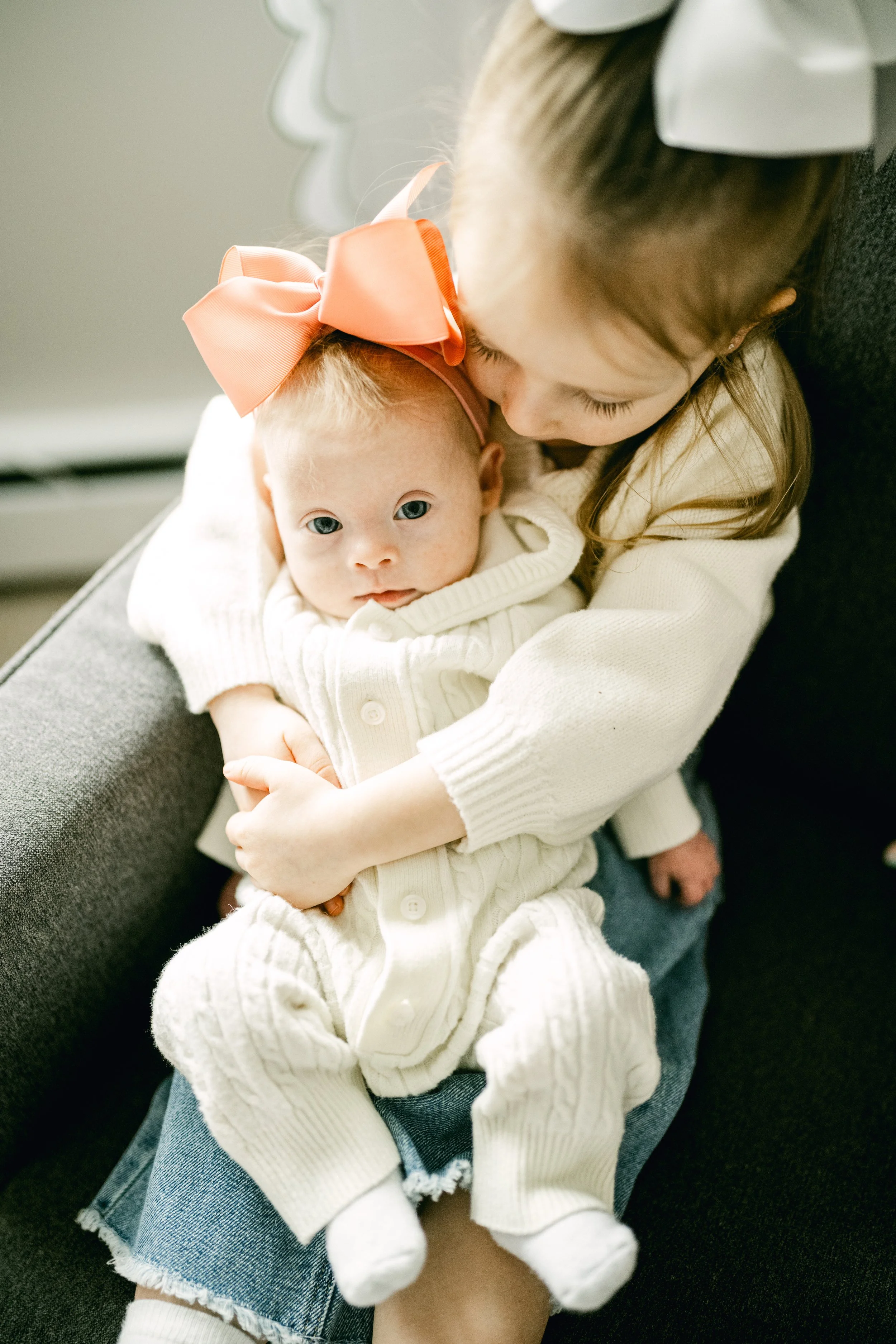 A young girl gently kisses a baby girl on her forehead while holding her in her arms. The baby is wearing a white knitted onesie and a large pink bow on her head.
