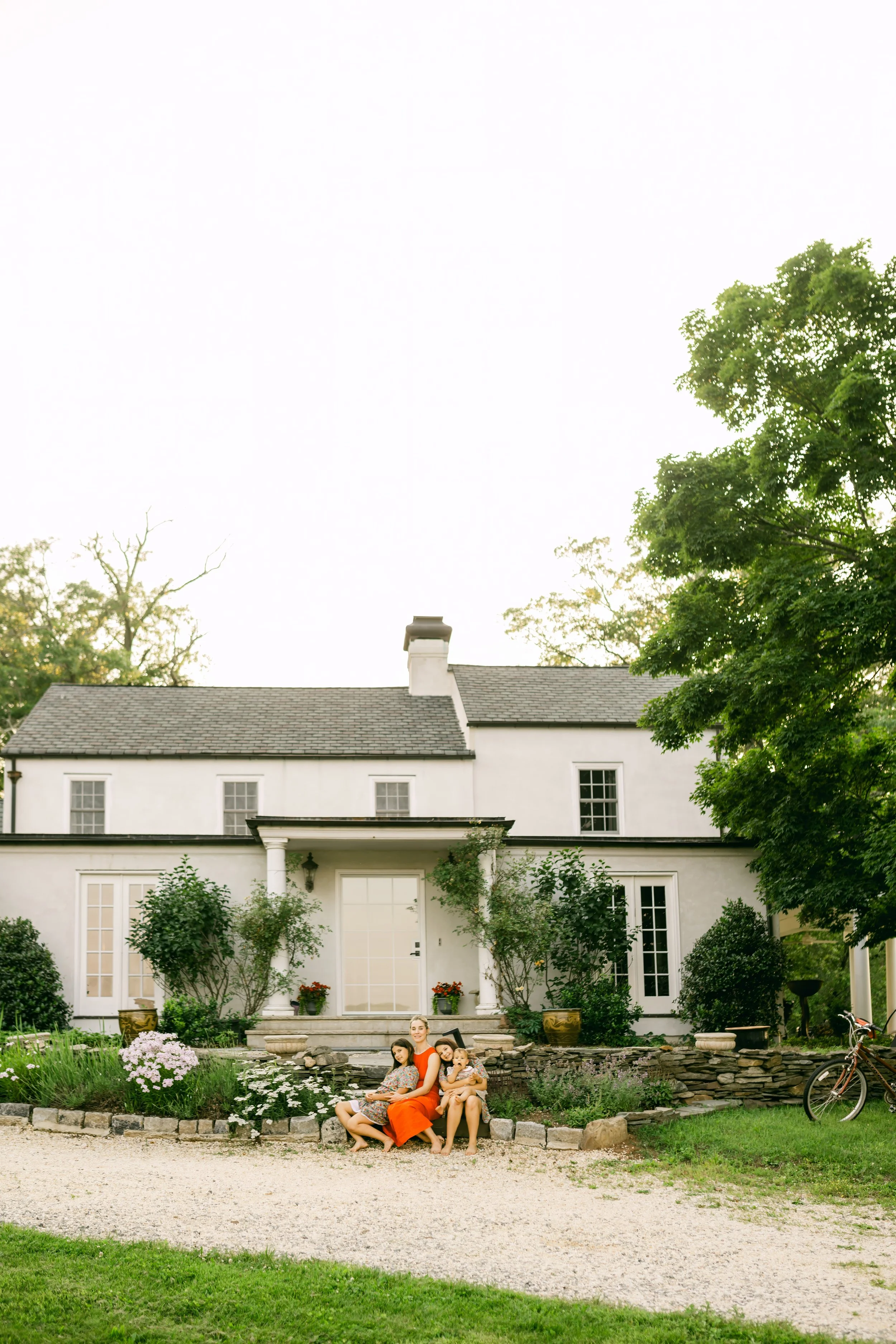 A woman and three children sitting on steps in front of a white house, surrounded by greenery and flowers, with a bicycle nearby.
