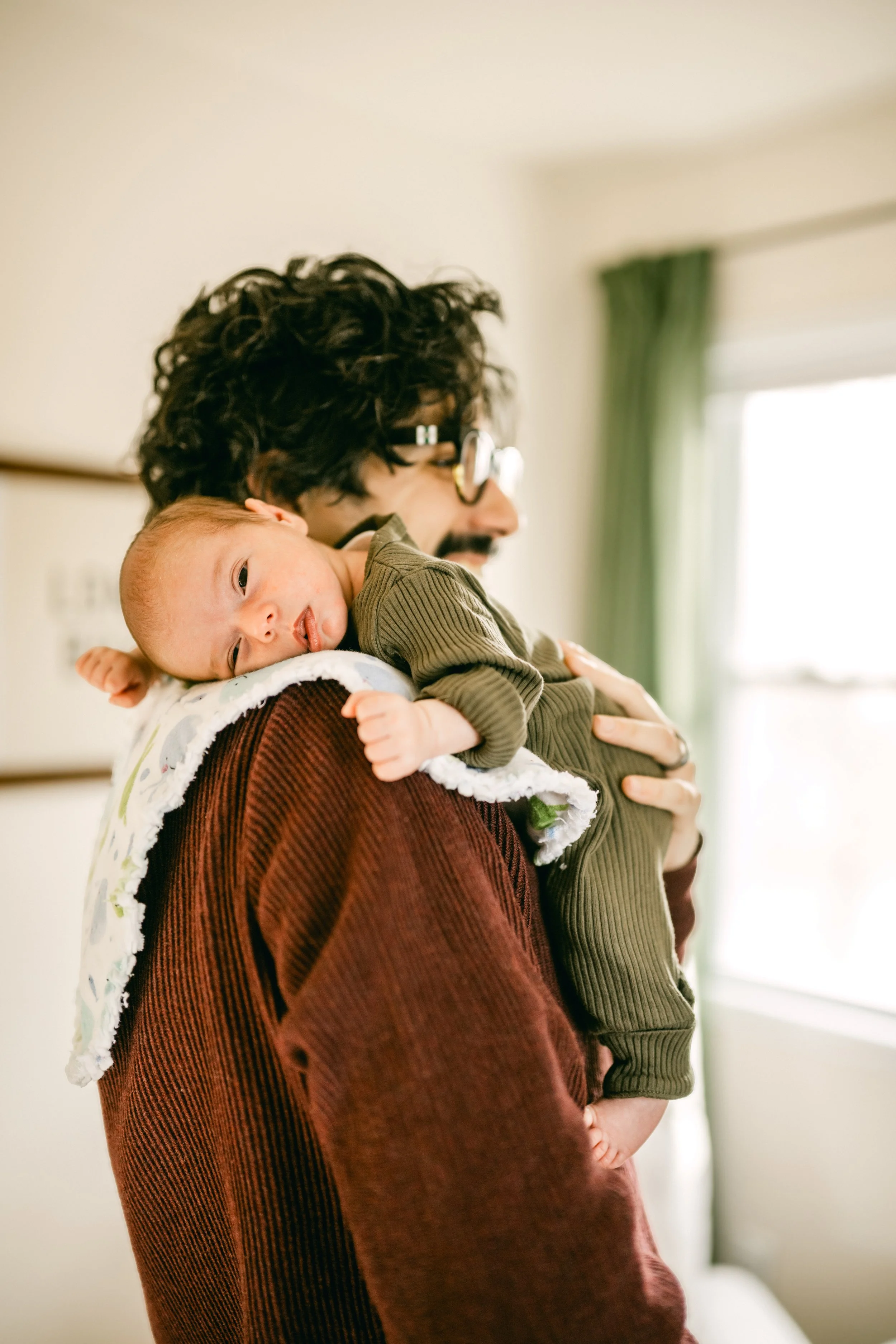 A man holding a baby on his shoulder indoors, with soft natural light coming through a window in the background.
