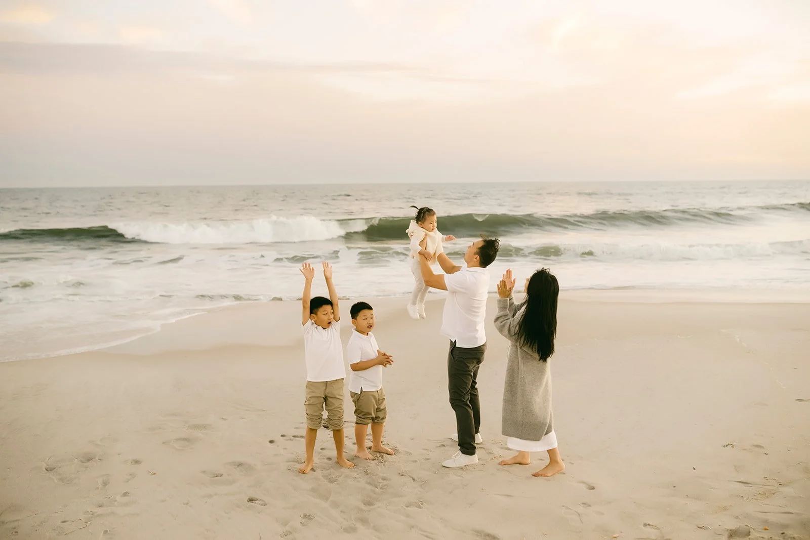 A family of five on the beach, with the father lifting a young girl into the air, two boys and a woman watching. The ocean and cloudy sky in the background.