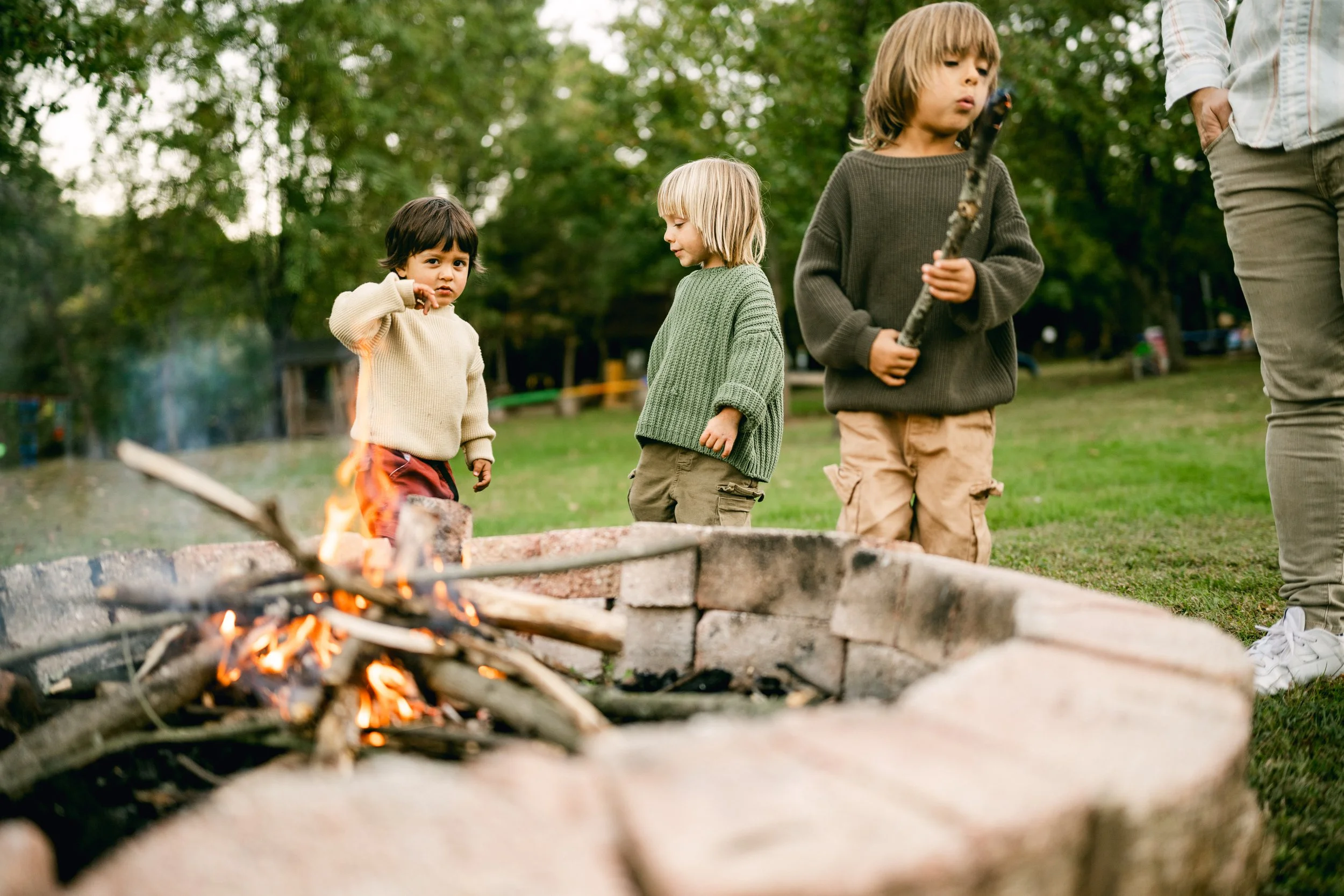 Three children standing by a campfire outdoors during daytime, with trees in the background.