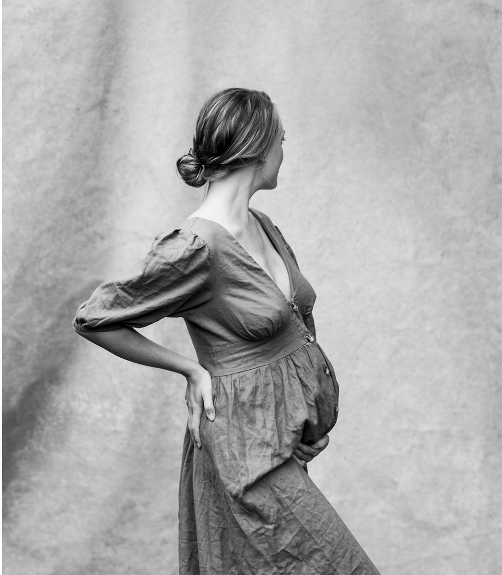 A pregnant woman with her hair in a bun, wearing a button-up dress, standing sideways and holding her belly with one hand while looking to the right.