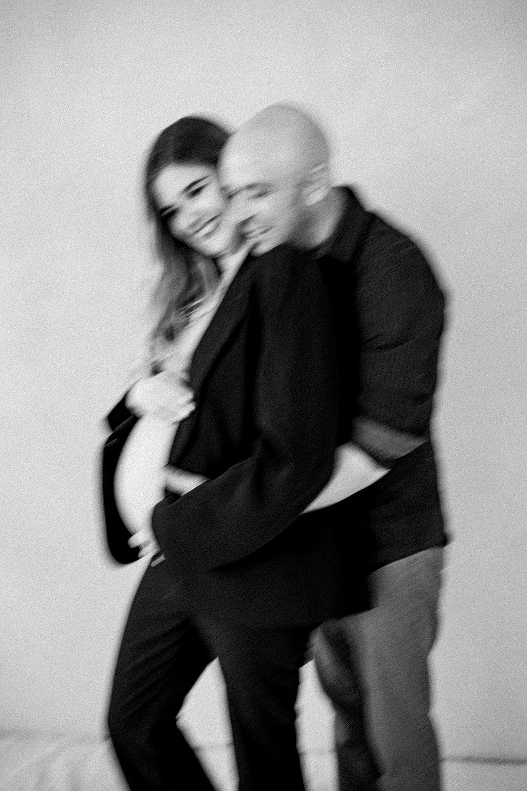 A blurred black-and-white photo of a smiling woman with long hair and a man with a shaved head, both in dark clothing, standing close together against a plain wall.