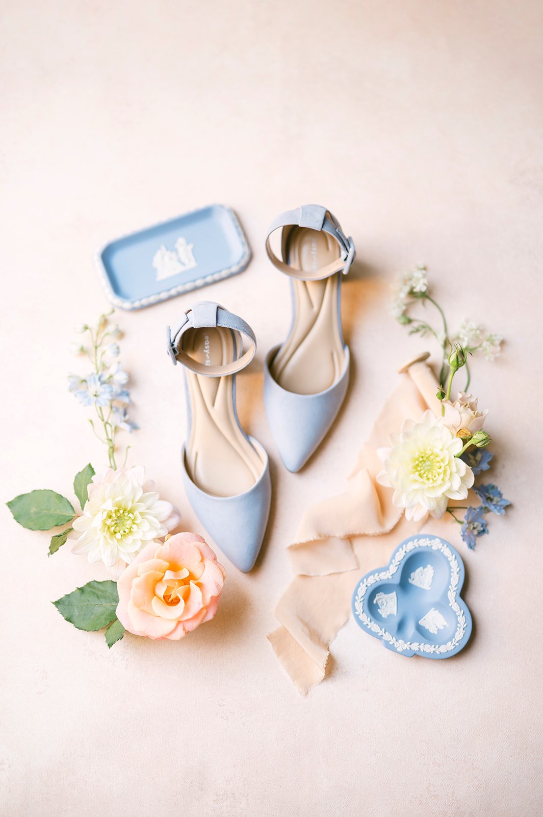 Light blue pointed-toe high heels with ankle straps, surrounded by pastel-colored flowers, vintage jewelry, and decorative items on a soft pink background.