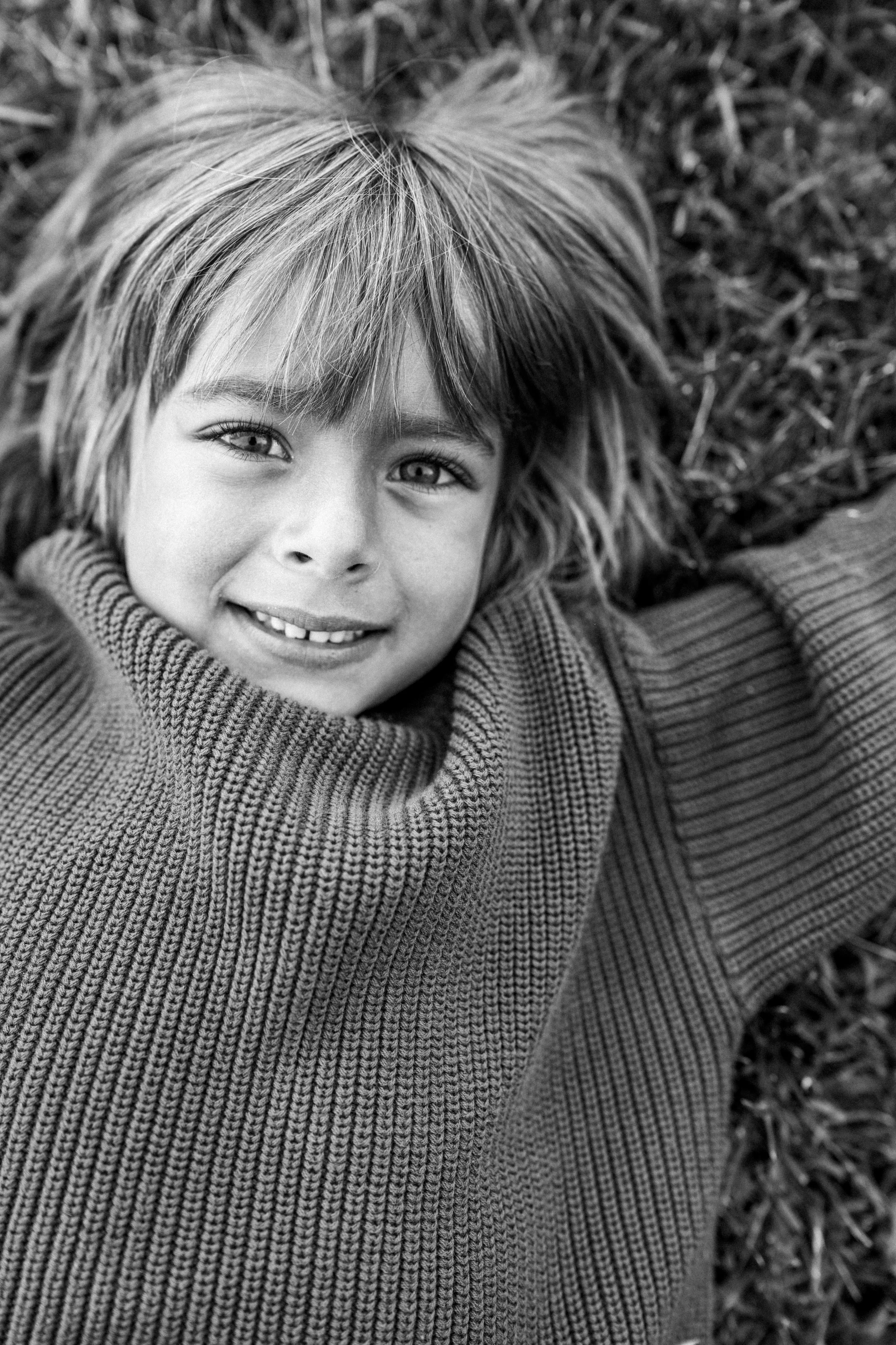 A happy young girl with long hair lying on the grass, smiling and looking up at the camera.
