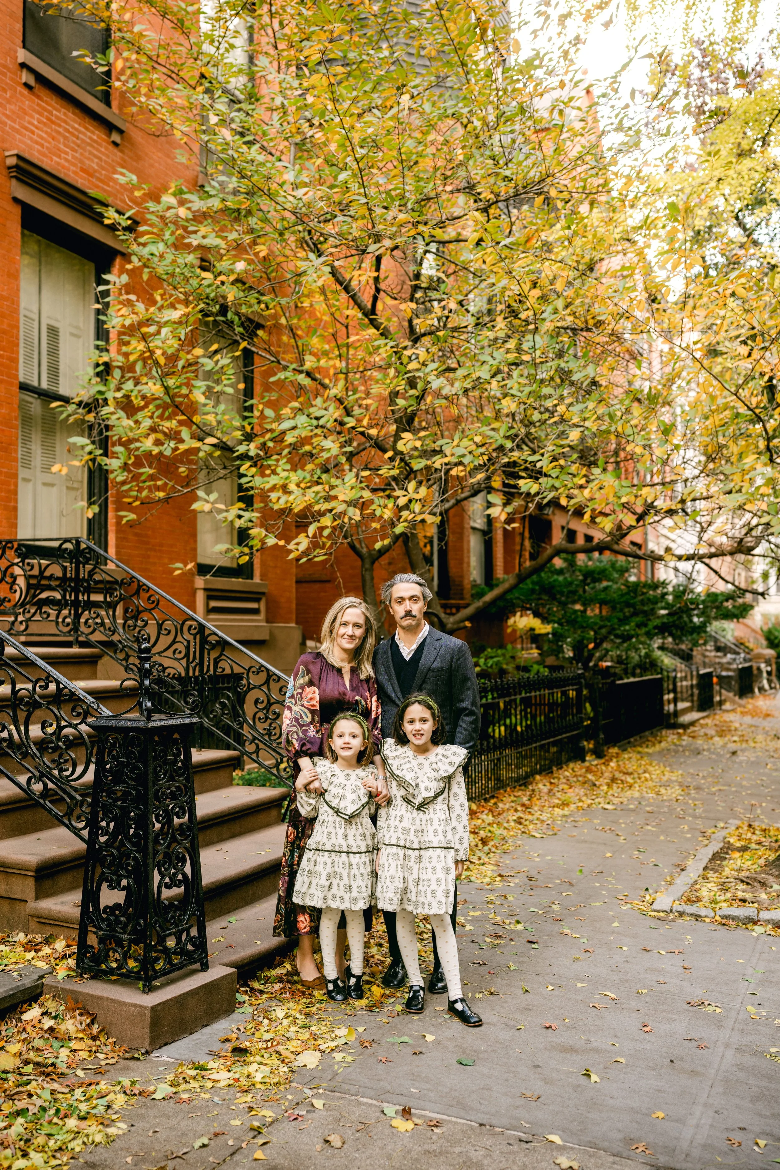 A family of four stands on the sidewalk in front of a brownstone with autumn leaves around them. The background features a large tree with yellow and orange leaves and a brick building with black wrought iron stairs.