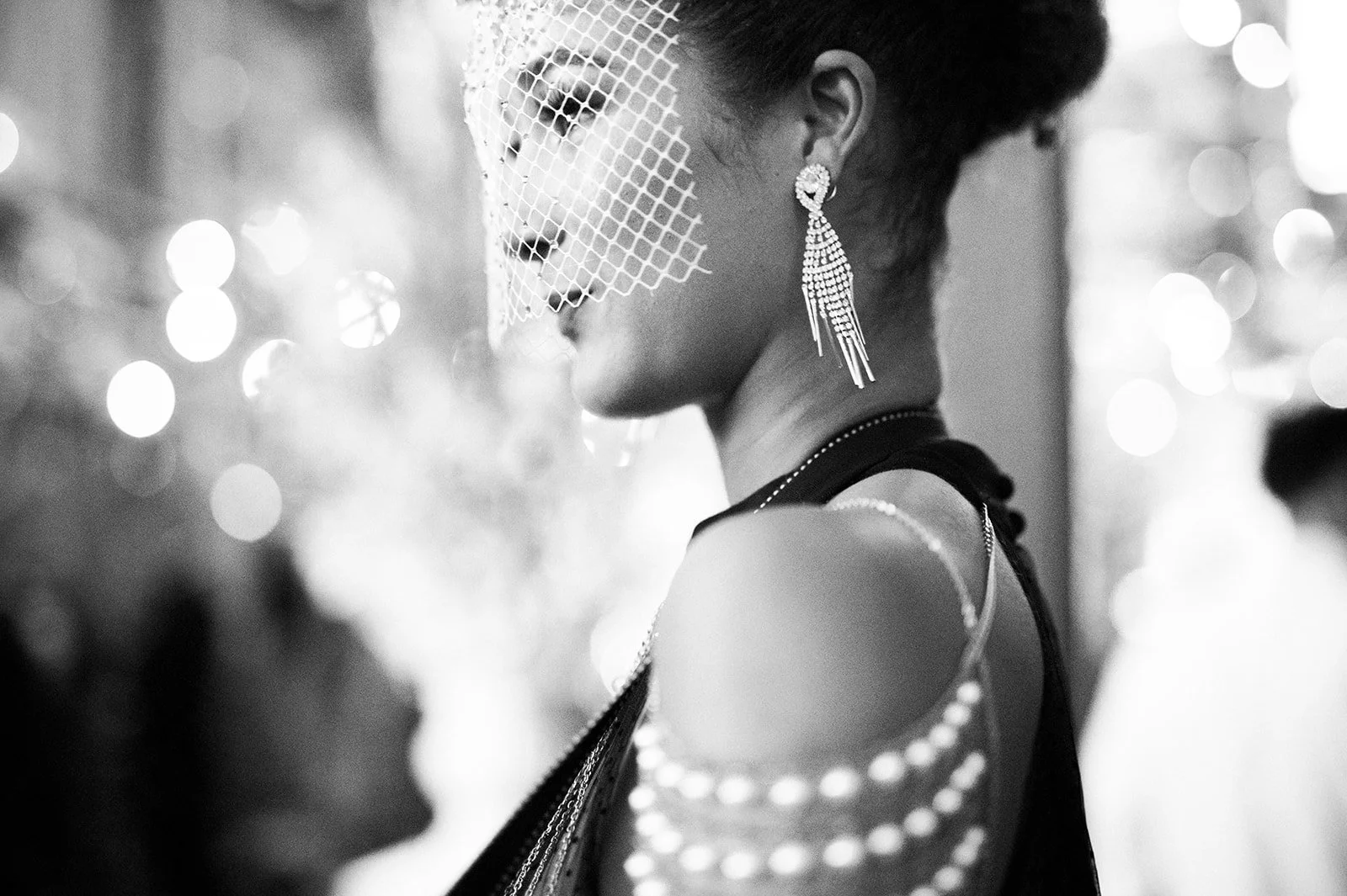 A woman in profile wearing a veil and ornate earrings at a formal event, with soft bokeh lighting in the background.
