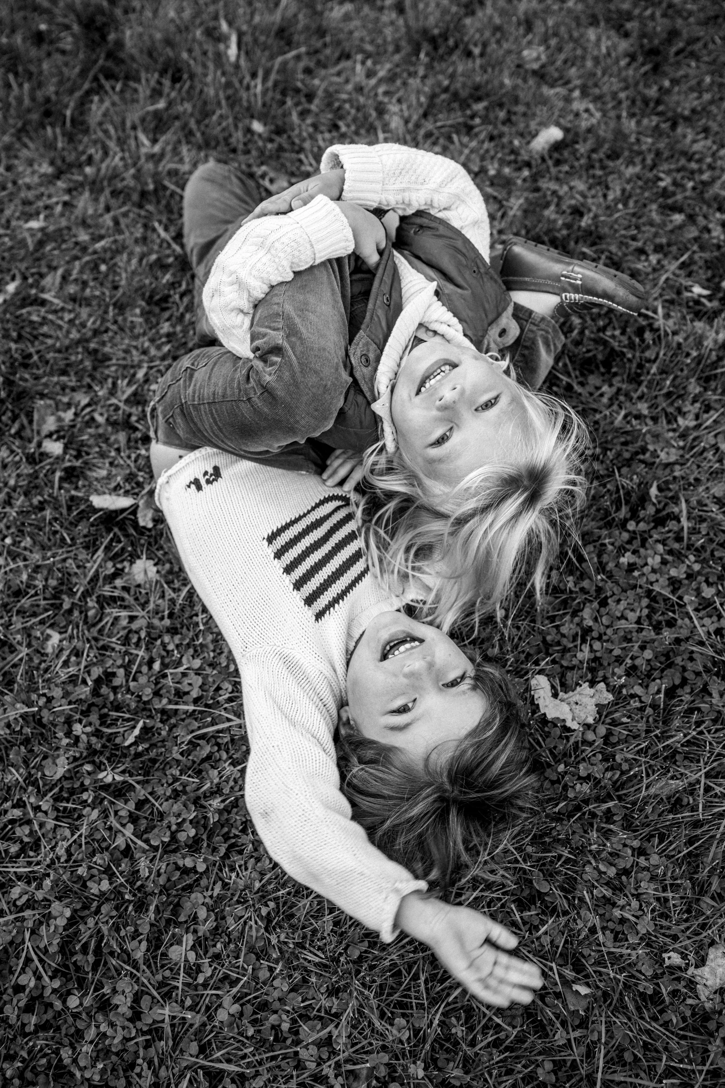 Two children lying on grass, smiling and looking up at the camera. One girl with long hair and the other girl with shorter hair.