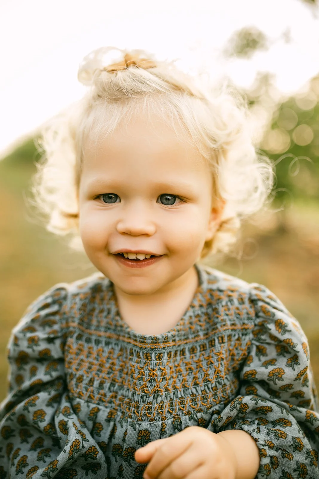Smiling young girl with curly blonde hair and blue eyes, wearing a gray dress with orange floral embroidery, outdoors with blurred greenery in the background.