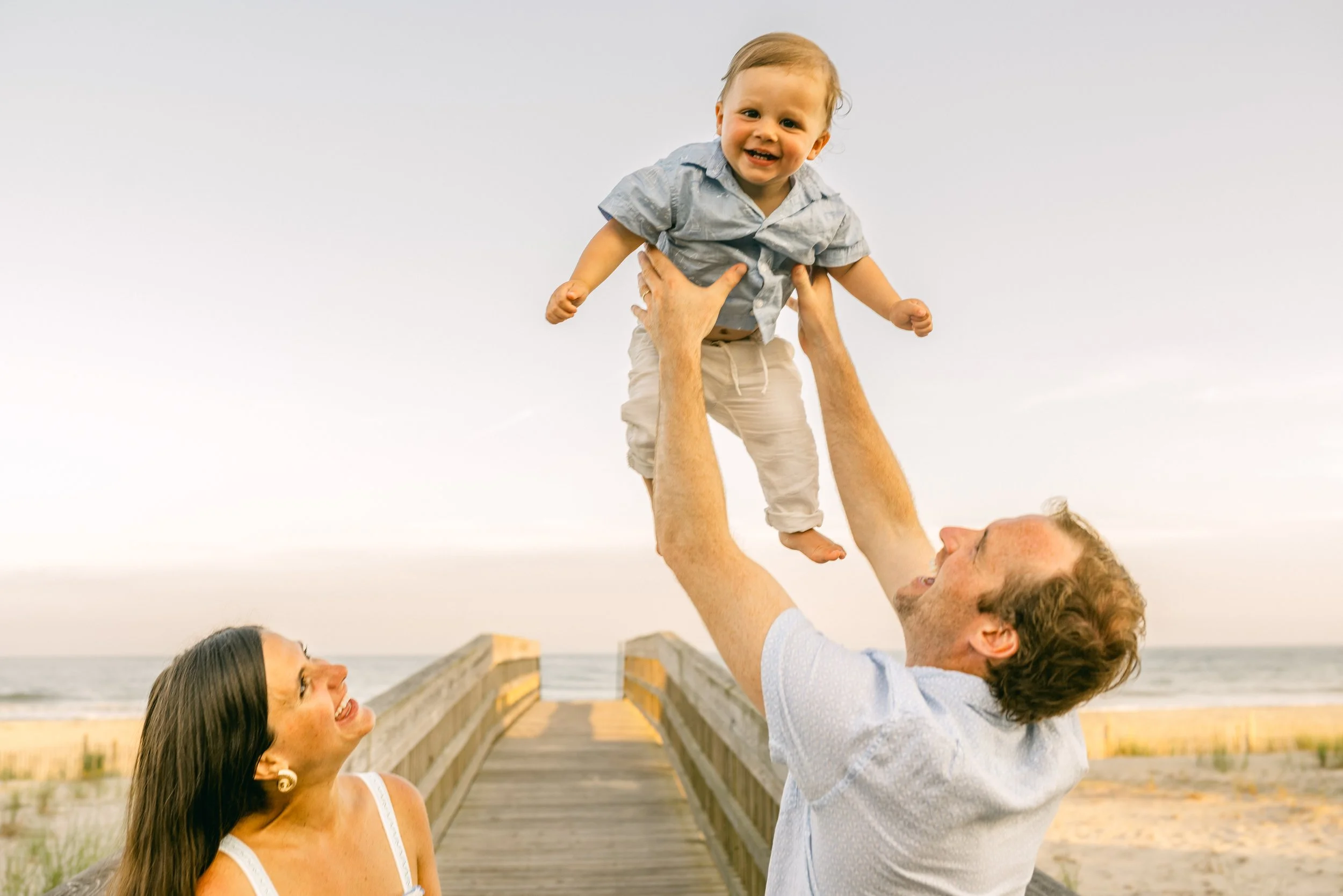 A man and woman playing with a young child on a beach pier at sunset, lifting the child in the air and smiling.