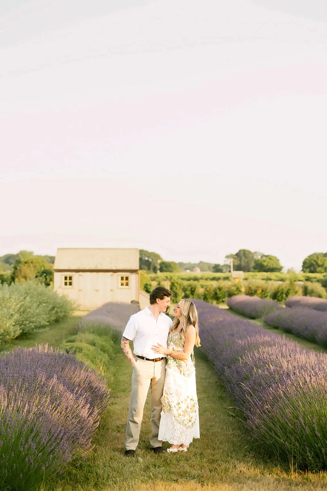 A couple stands close together on a grassy path between rows of blooming lavender, with a rustic wooden barn in the background, under a pastel sky.