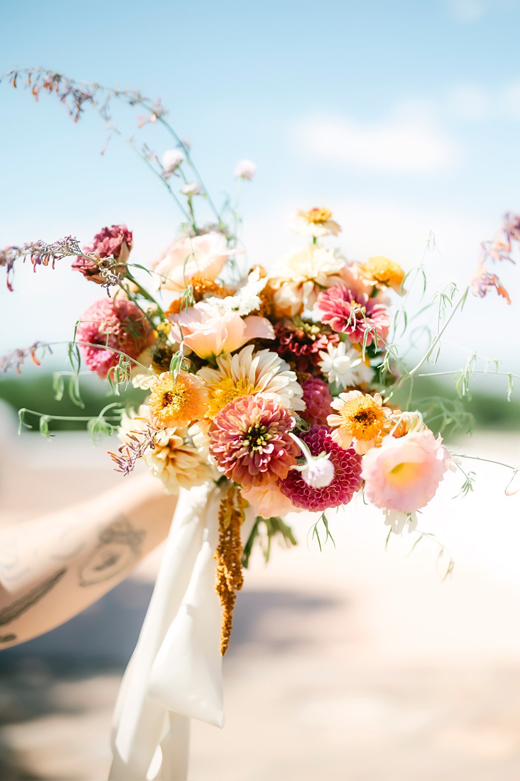 Close-up of a colorful bouquet of flowers held outdoors against a bright sky, featuring pink, white, yellow, and orange blooms with green foliage.