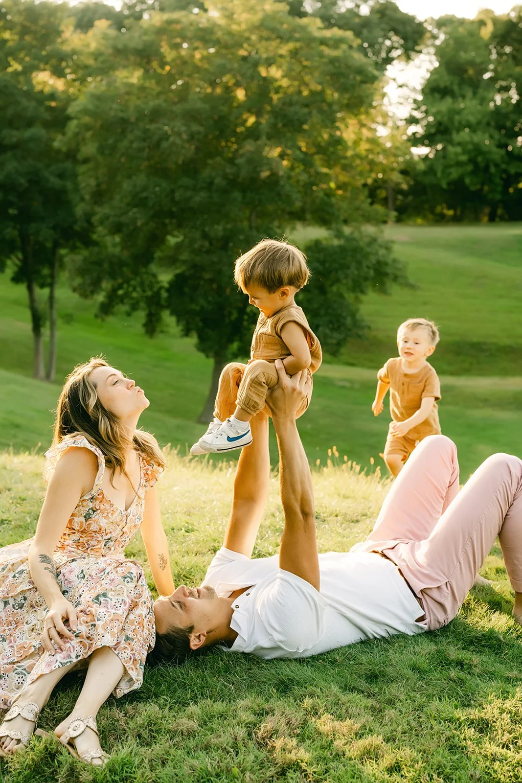 Family playing in a park with lush green trees and grass, with a young woman, a man lying on the ground, and two small children, one being lifted by the man.
