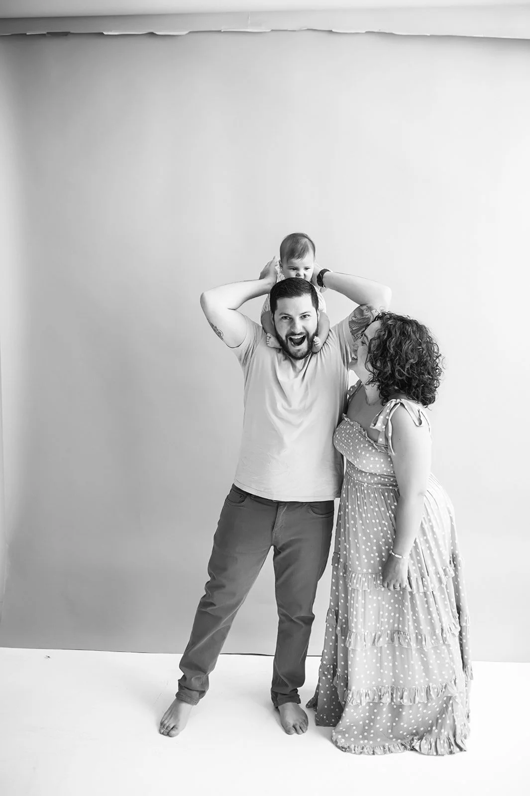 A happy family of three, including a man, woman, and baby, standing against a blank background. The man is barefoot, wearing a t-shirt and pants, and holding the baby on his shoulders. The woman is wearing a long, polka-dot dress and leaning in to ki