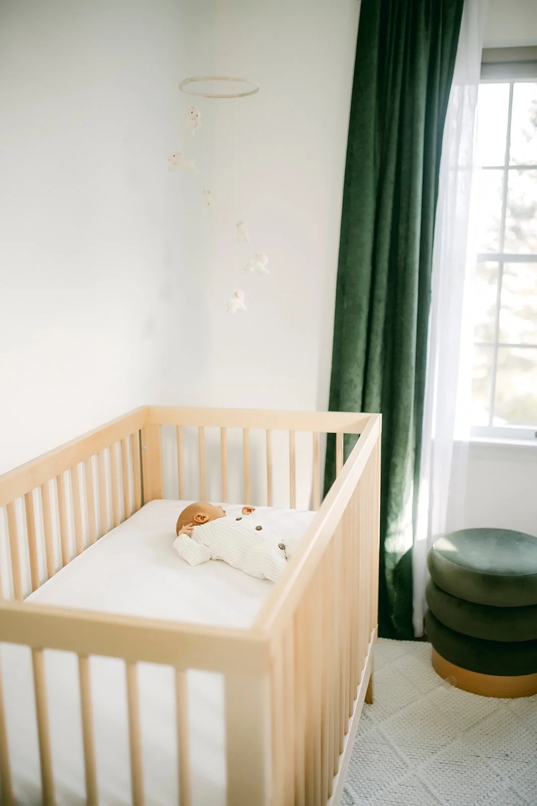 A nursery with a light wooden crib, a doll dressed in white lying inside, a grouped set of green cushions, and dark green curtains by a window.