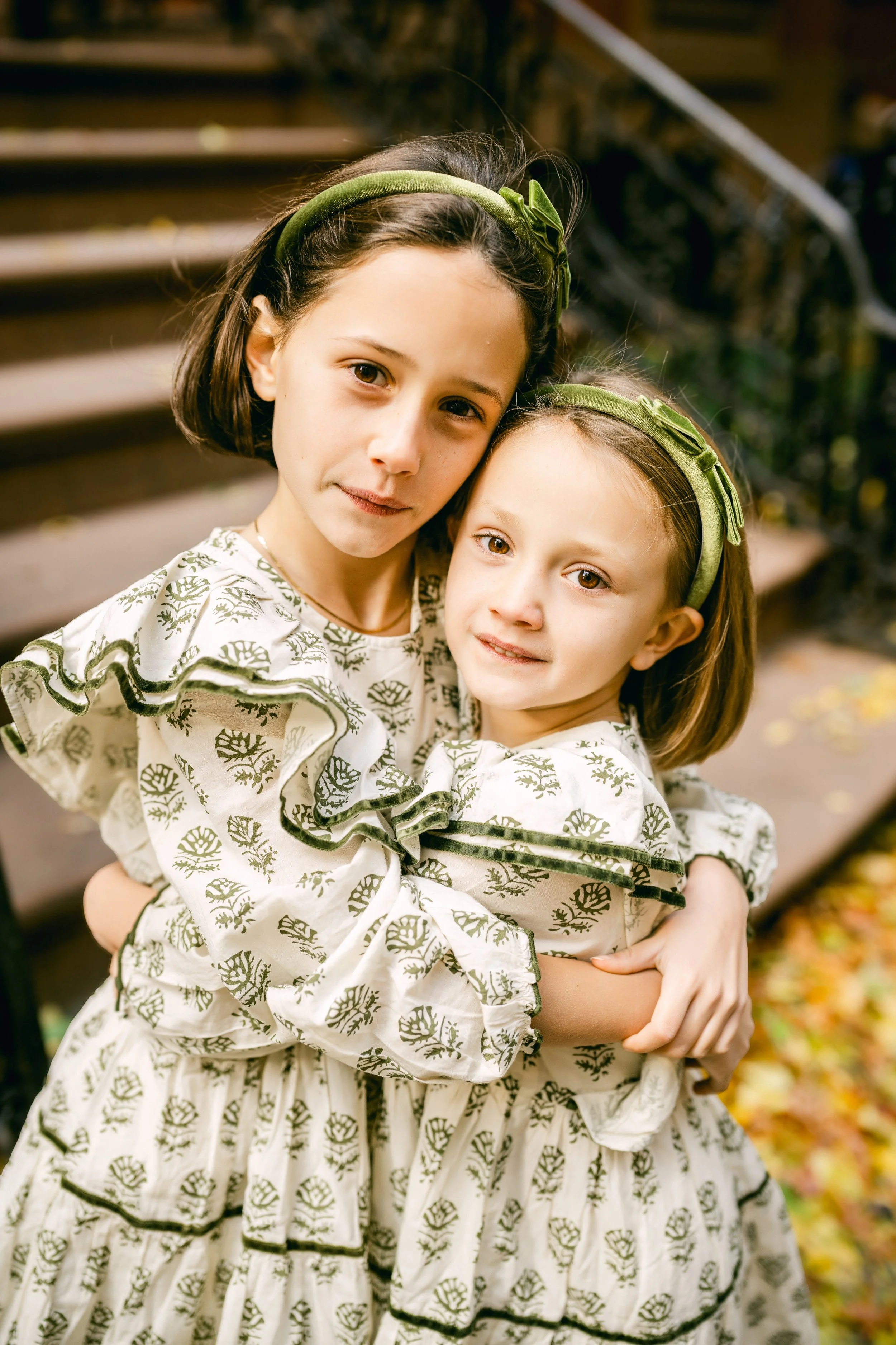 Two young girls hugging each other outdoors on a staircase, wearing matching white dresses with green leaf patterns and green headbands, with autumn leaves on the ground.