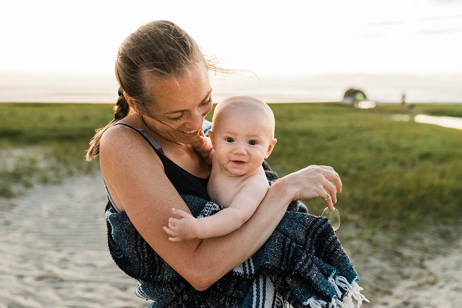 A woman holding a baby at the beach during sunset, both smiling and wrapped in a towel.