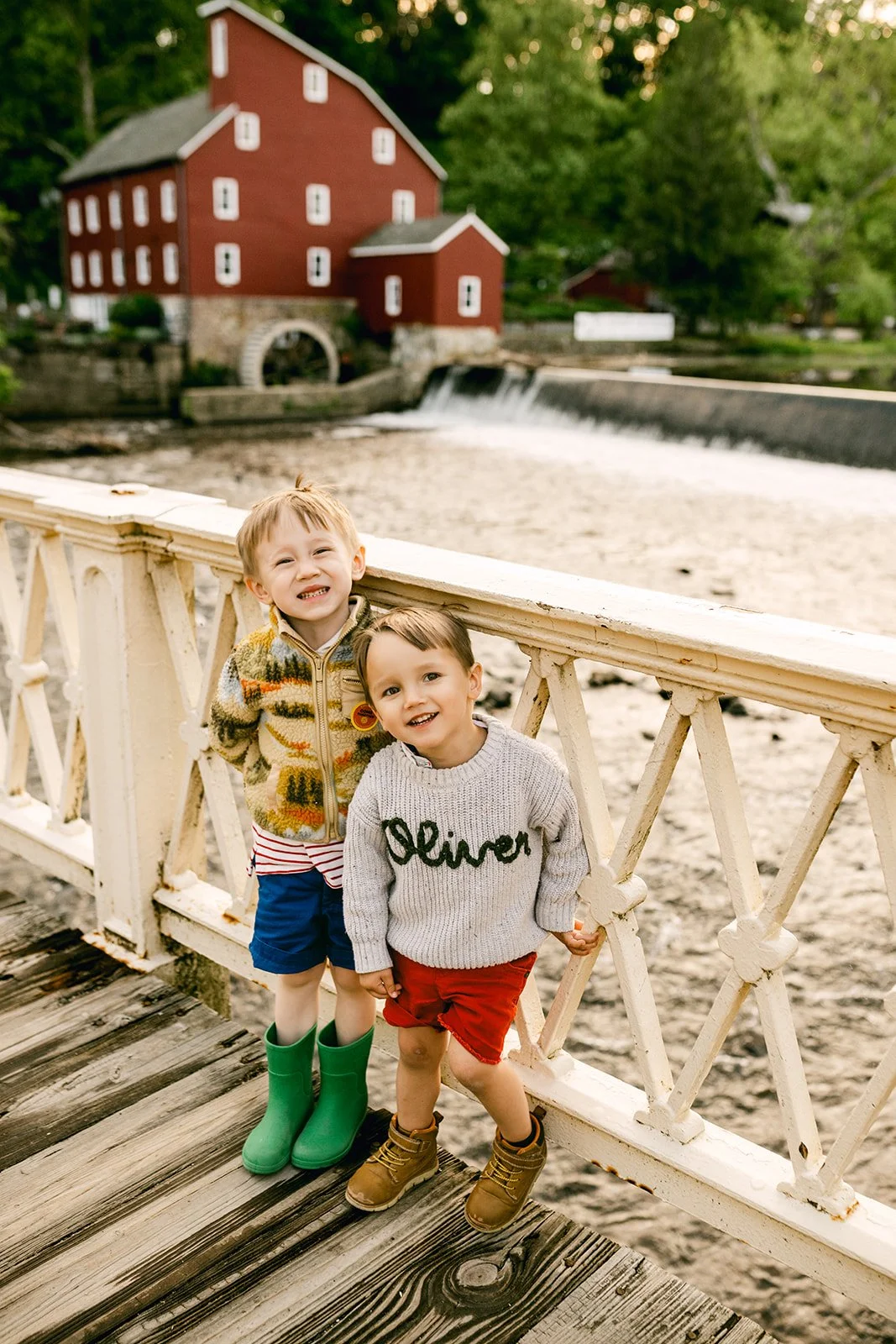 Two young boys standing on a wooden bridge with a red mill in the background, smiling and posing for a photo.