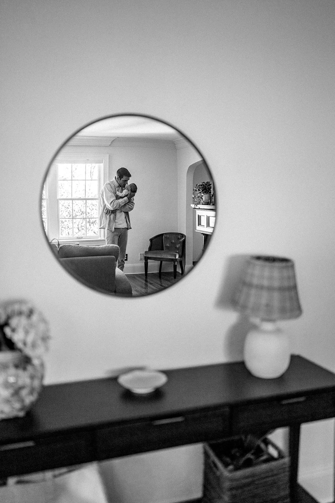 A black-and-white photo showing a man holding a baby seen through a circular mirror on a wall, with a table and a lamp in the foreground.