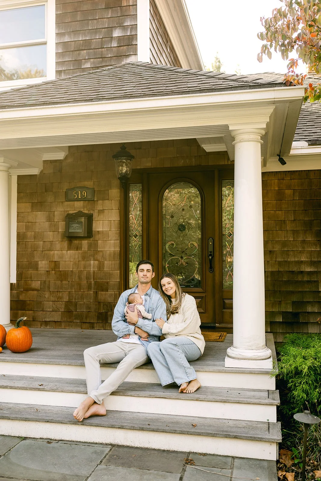 A family of three sitting on the front porch of a house with pumpkins nearby, smiling at the camera.