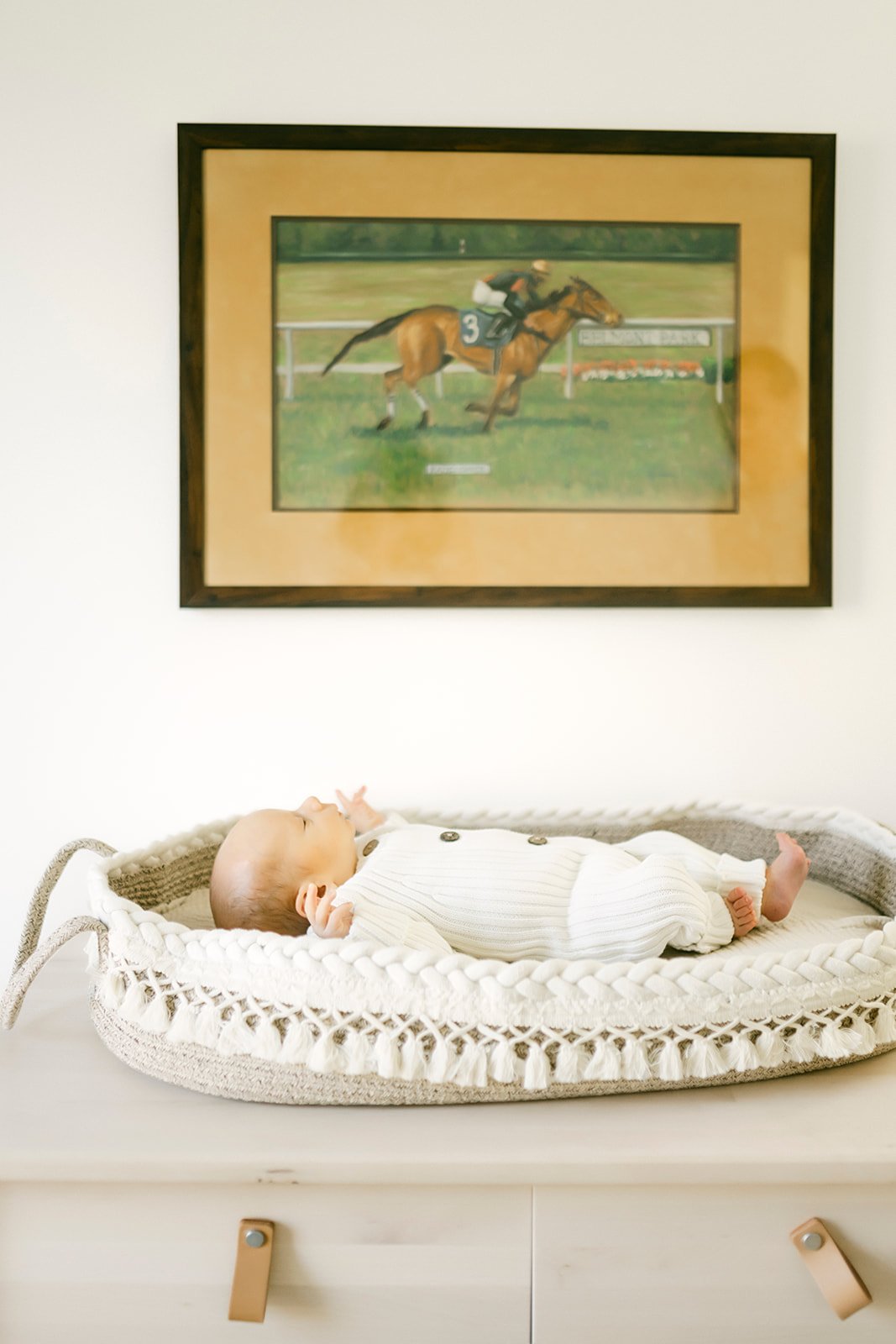 A baby lying on a crocheted baby bed in front of a wall with a framed picture of a horse race.