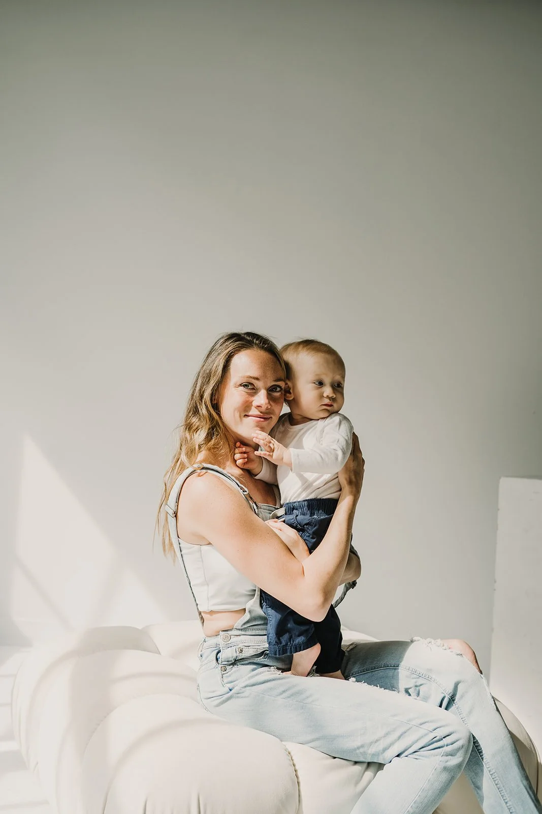 A woman sitting on a white sofa holding a young boy in her arms, both looking towards the camera, with a plain light-colored background.