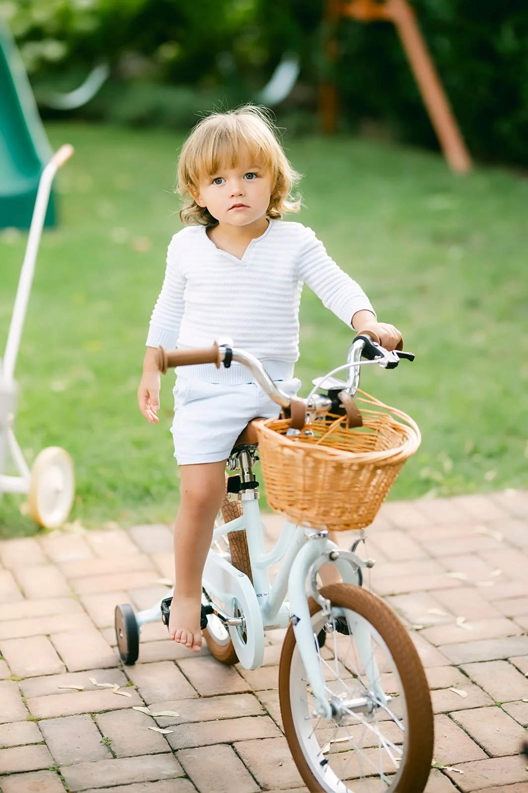 A young child with blonde hair and blue eyes riding a white bicycle with a wicker basket on the front in a backyard, with a green lawn and a slide visible in the background.