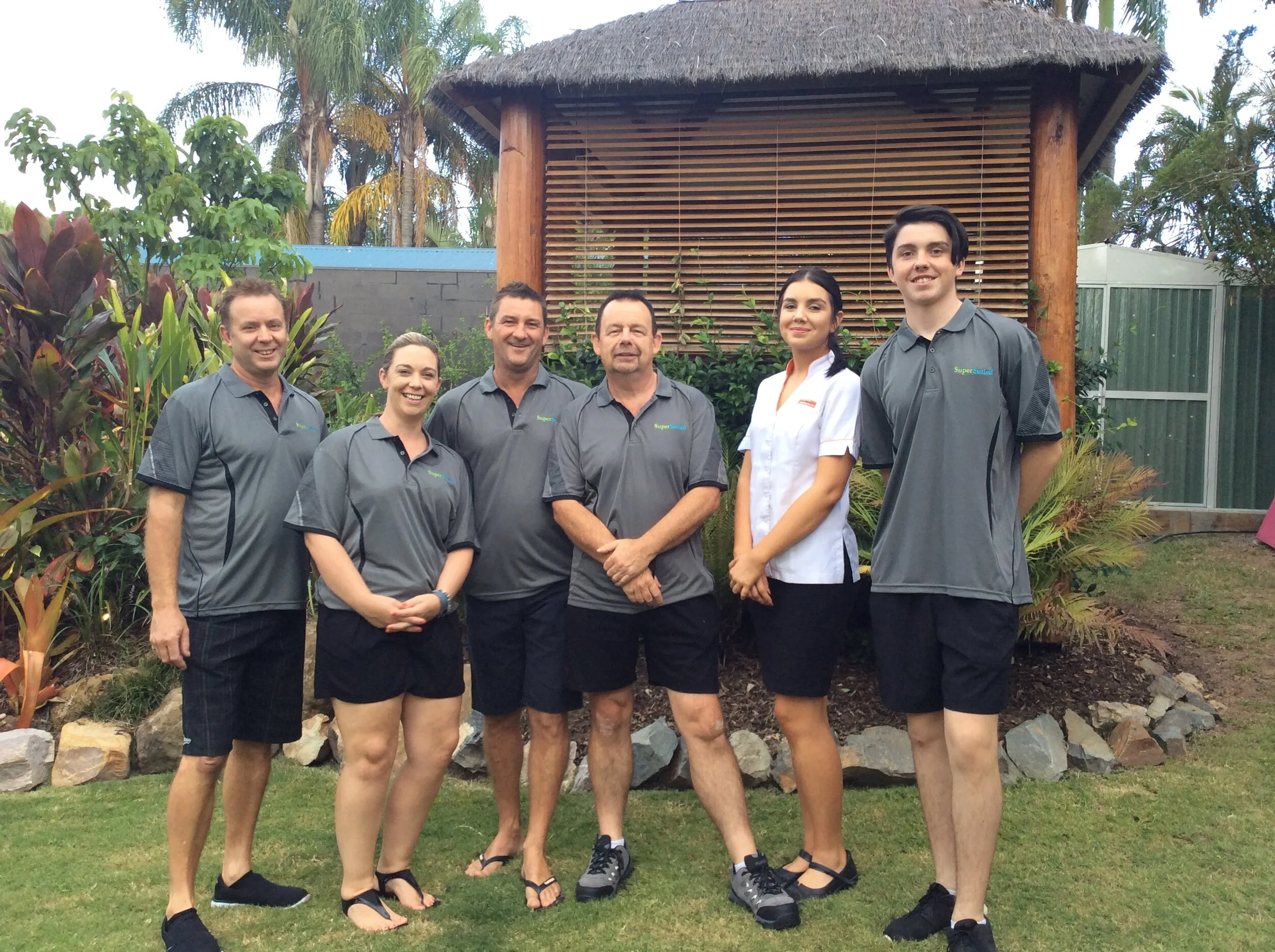 Group of six people standing outdoors in front of a wooden structure, wearing uniforms, smiling, surrounded by greenery and tropical plants.