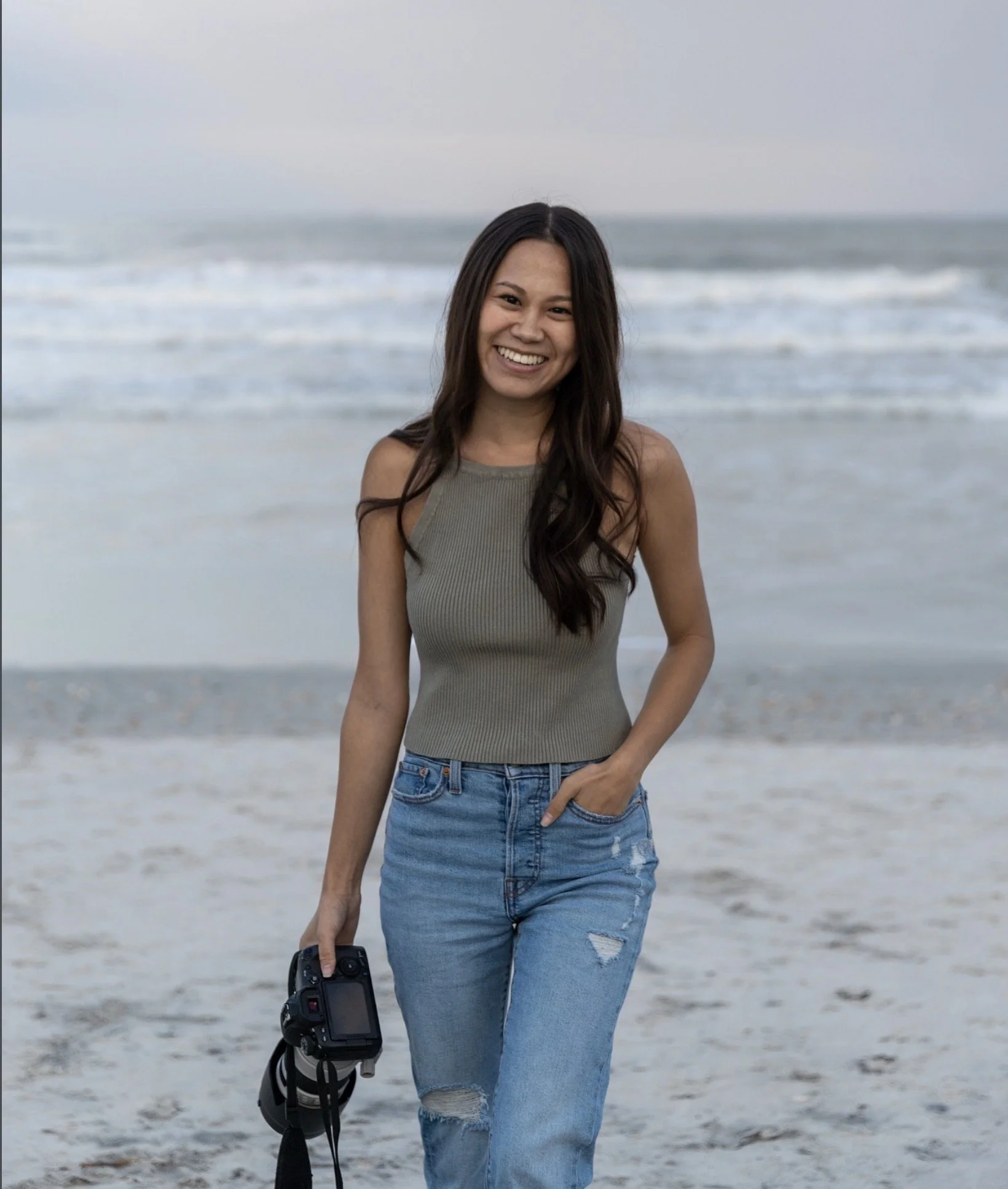 A woman with long dark hair smiling at the beach while holding a camera in her left hand and wearing a sleeveless beige top and ripped blue jeans