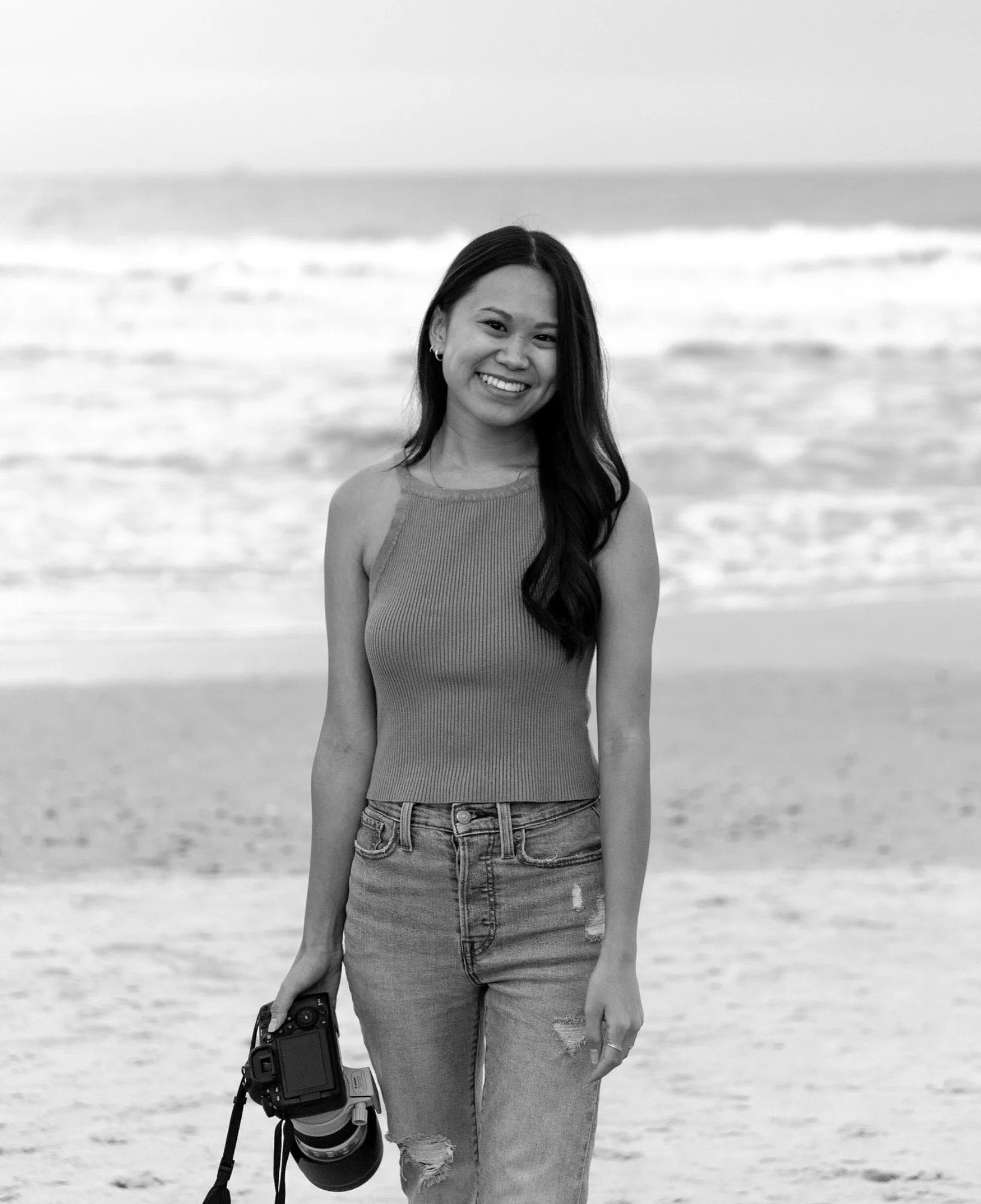 A woman smiling and holding a camera with a strap at the beach, in black and white.