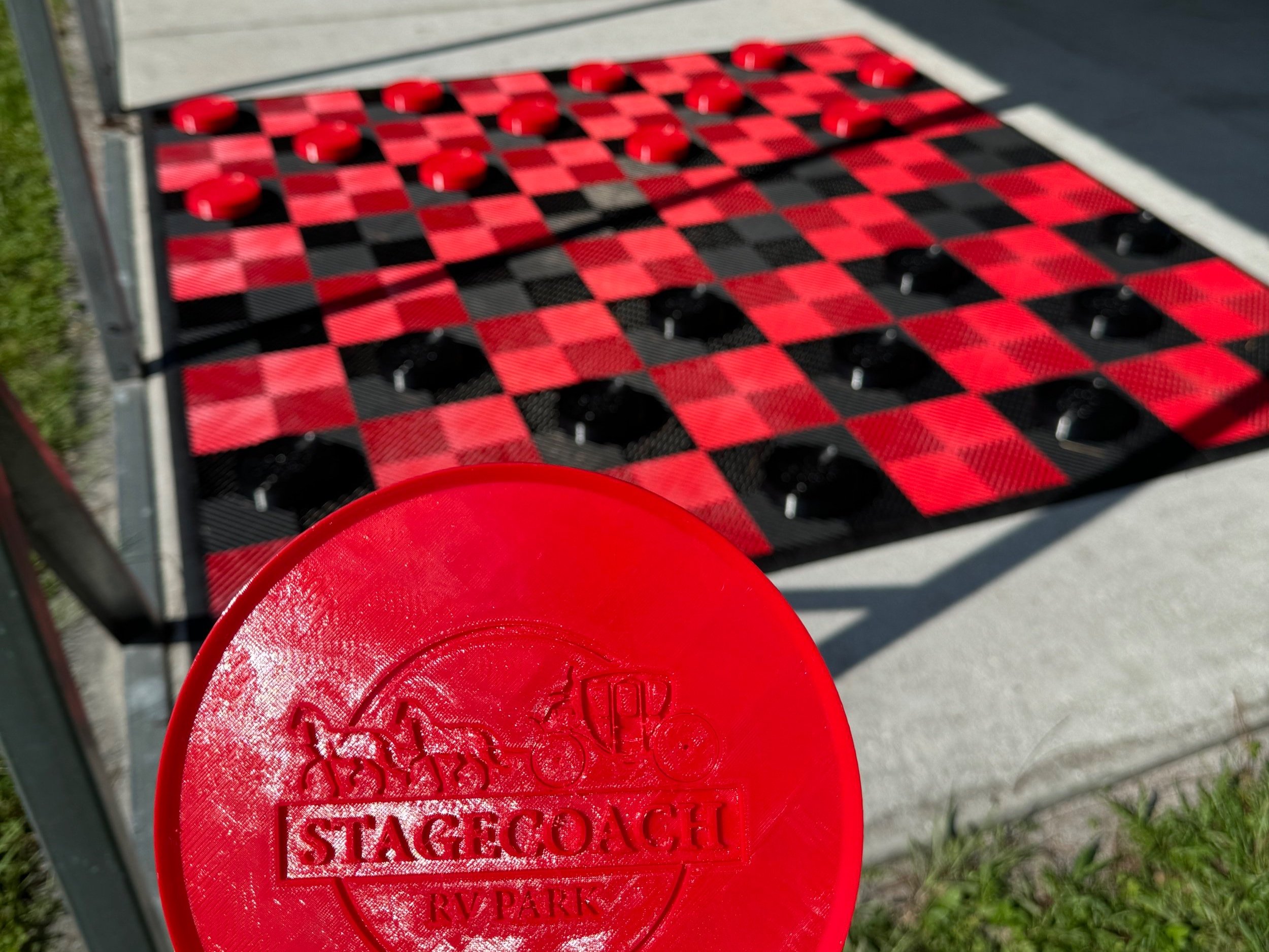 A red checker game disc with the Stagecoach RV Park logo, in front of a large outdoor checkers game with red and black checkered squares and round black pieces.