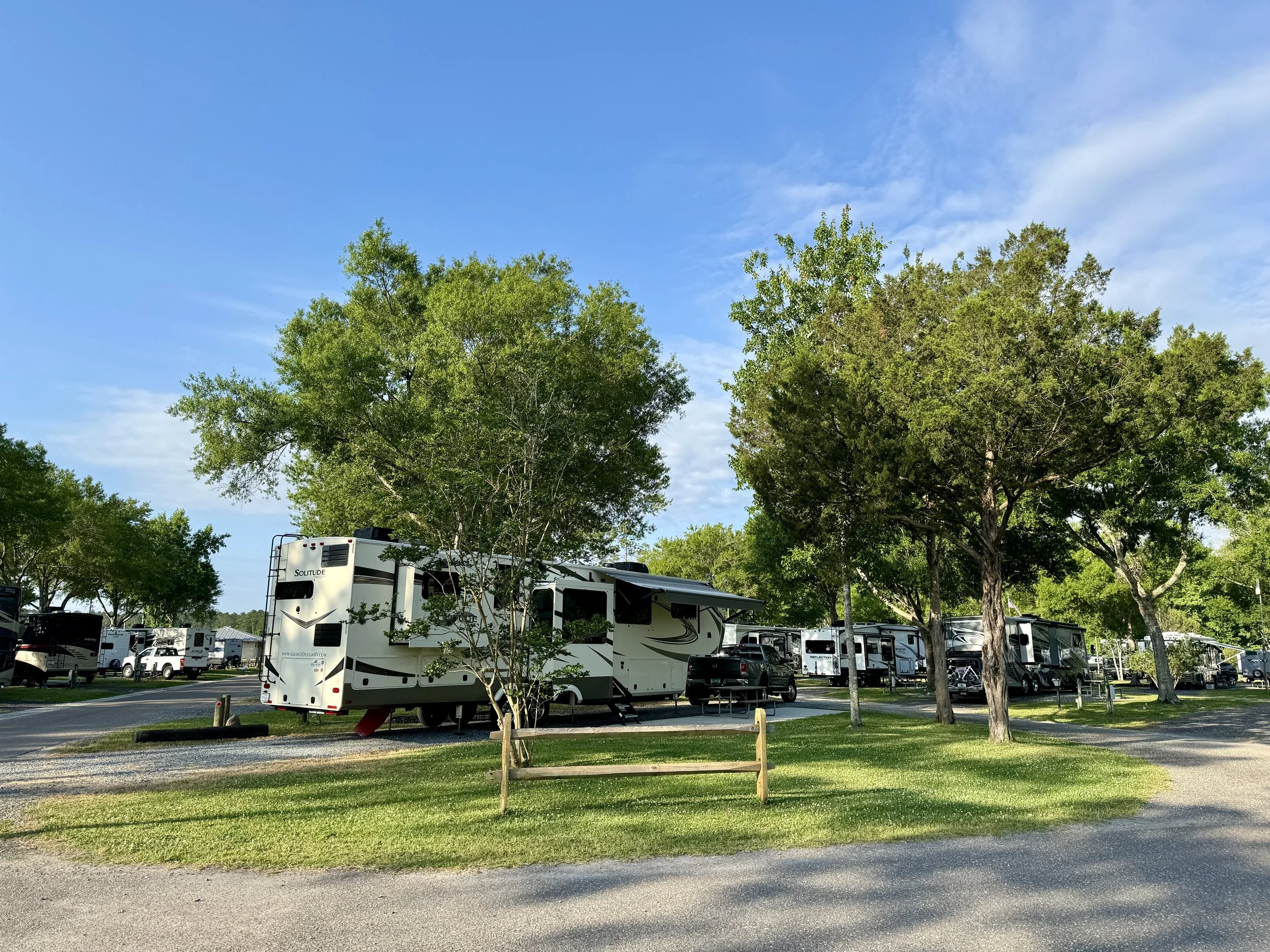 RV campground with multiple fifth-wheel RVs surrounded by green trees and a grassy area under a blue sky with some clouds.